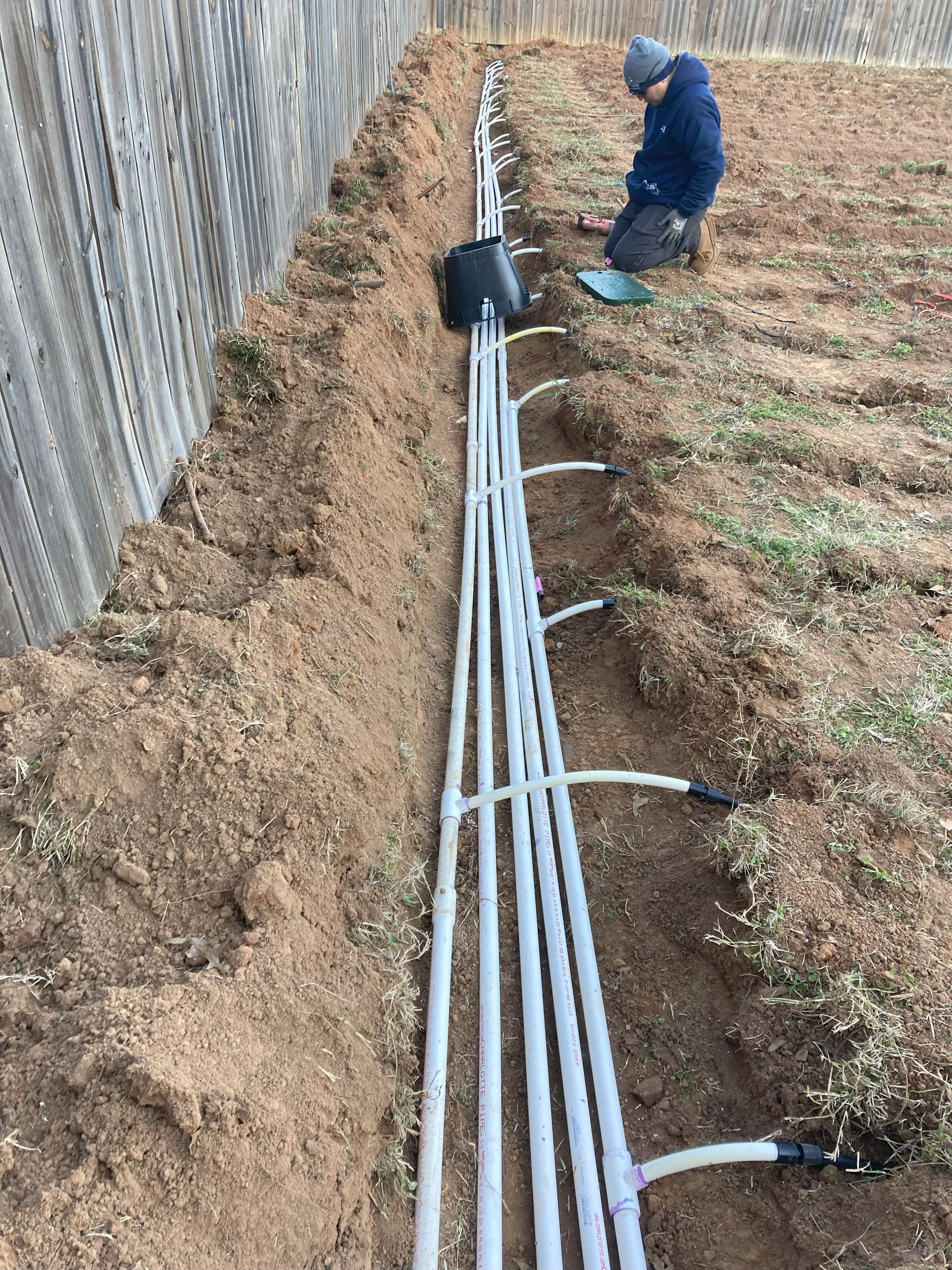A man is kneeling in the dirt next to a bunch of pipes.