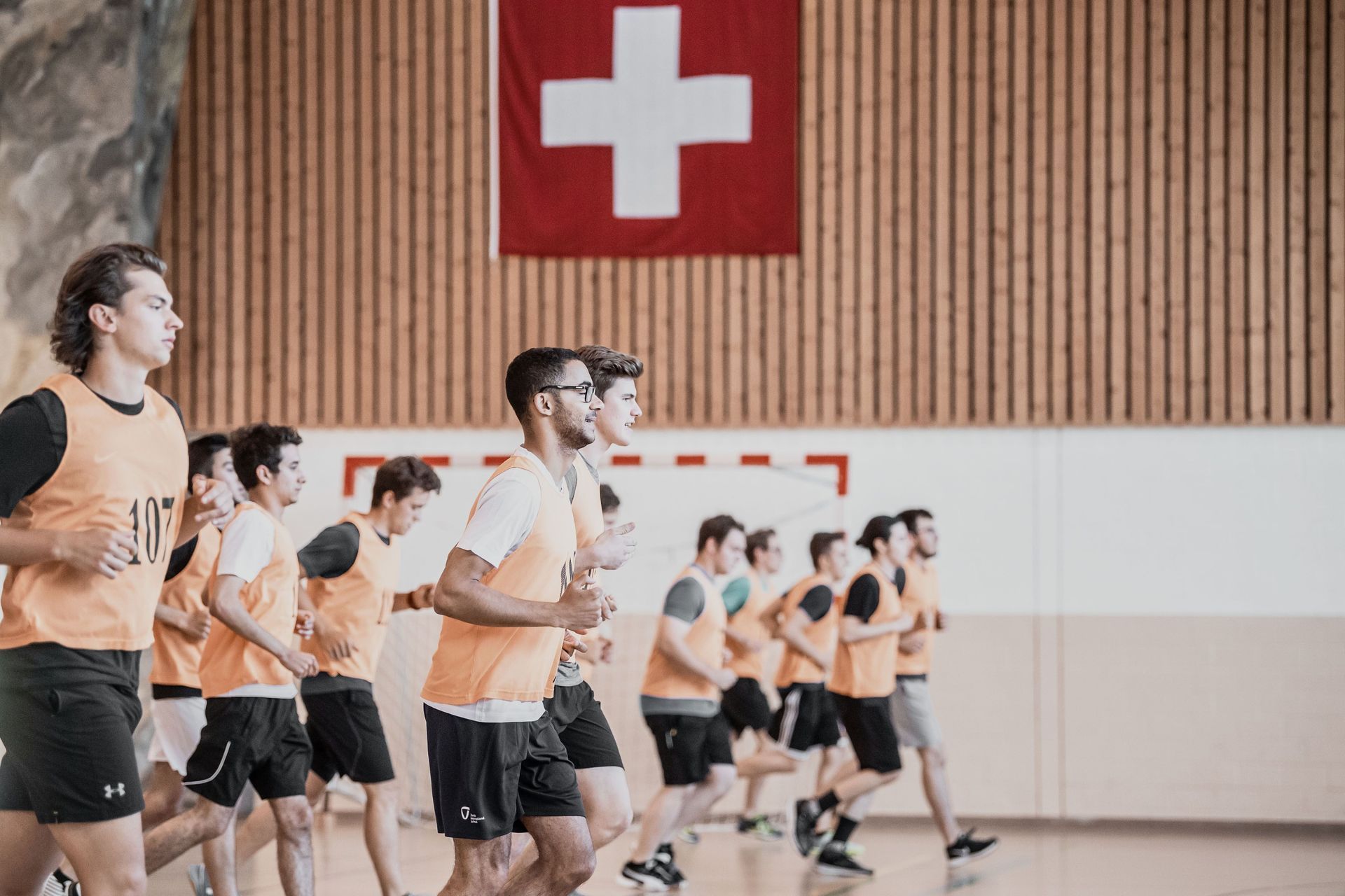 Eine Gruppe von Menschen in orangefarbenen T-Shirts trainiert in einem Fitnessstudio unter einer Schweizer Flagge.