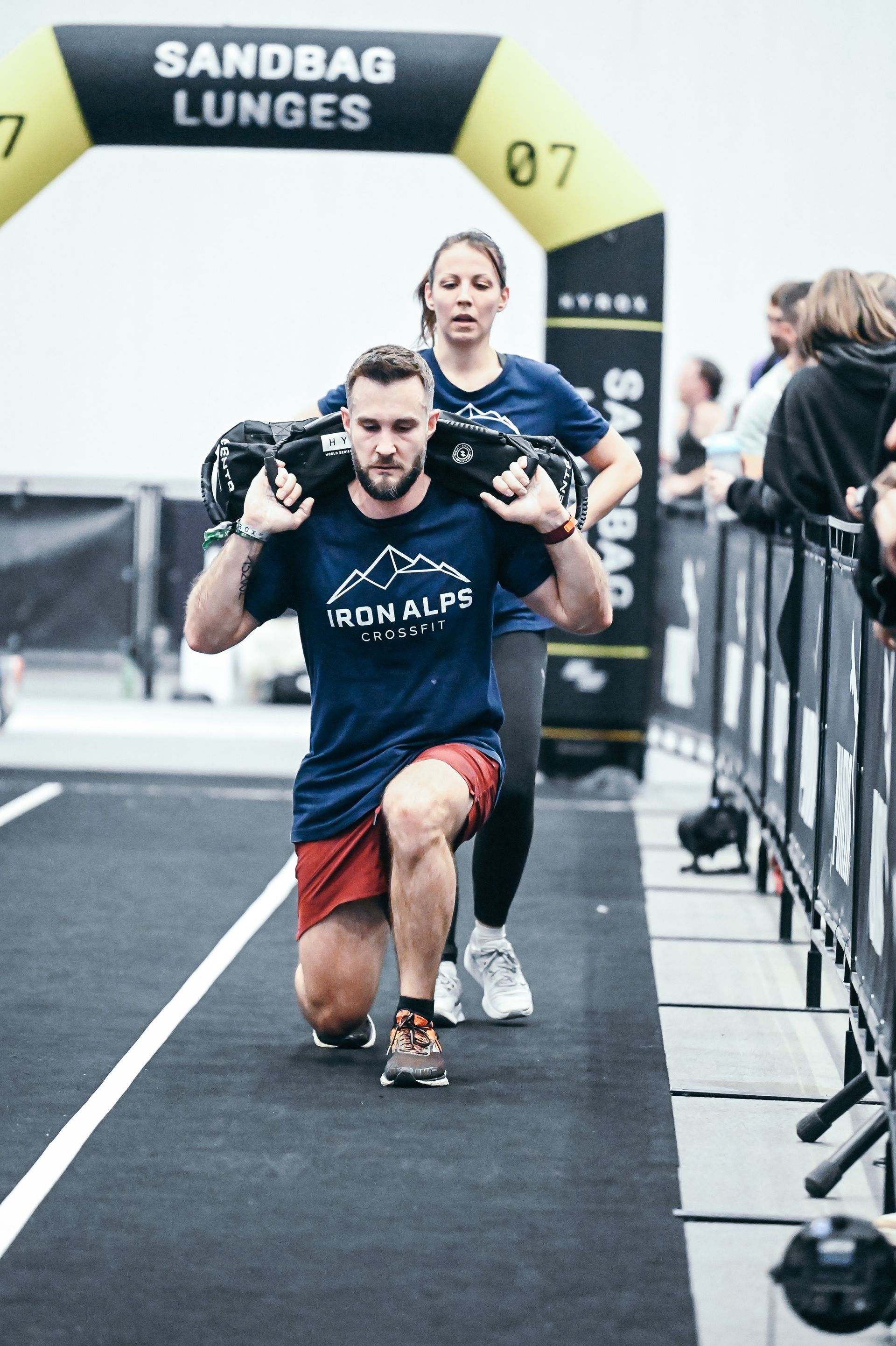 Zwei Athleten absolvieren bei einer Fitnessveranstaltung einen Hindernisparcours mit Sandsäcken.