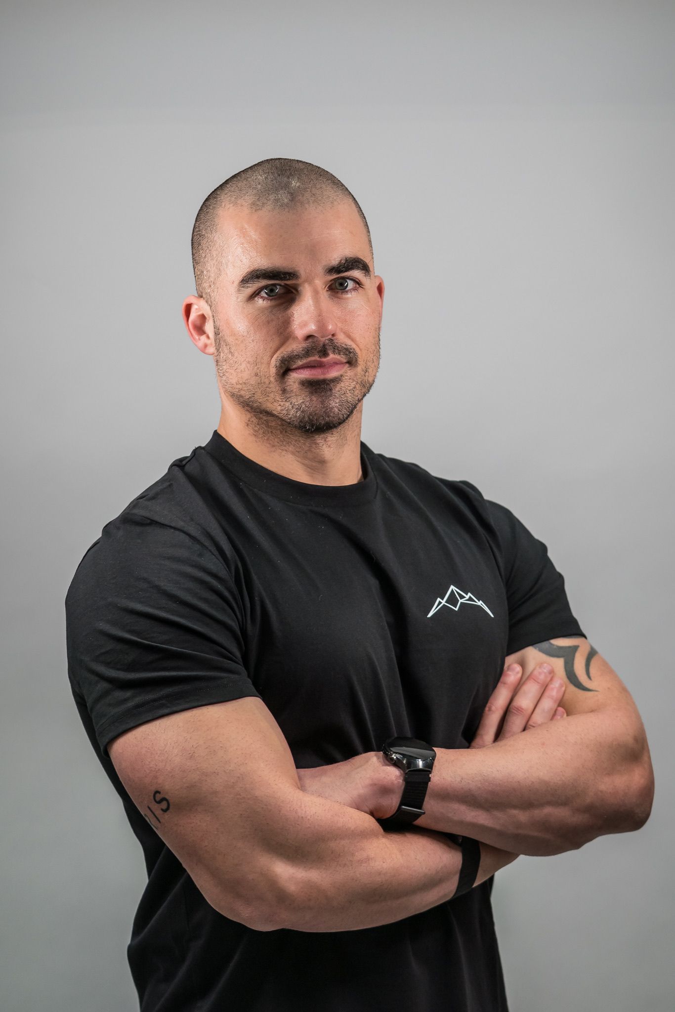 Ein Mann mit verschränkten Armen und einem schwarzen T-Shirt steht vor einem grauen Hintergrund.