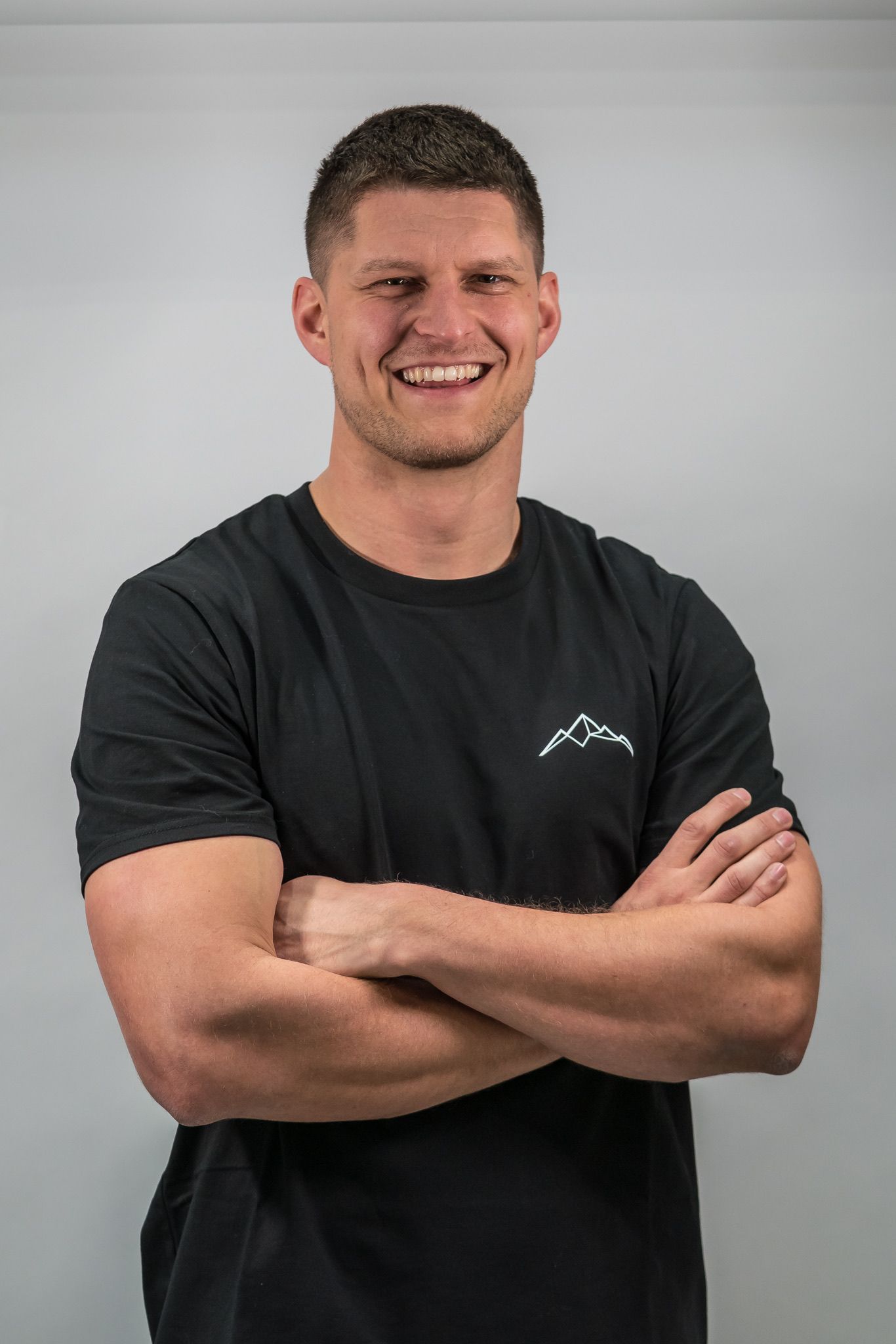 Lächelnder Mann in schwarzem T-Shirt mit verschränkten Armen vor einfarbigem grauen Hintergrund
