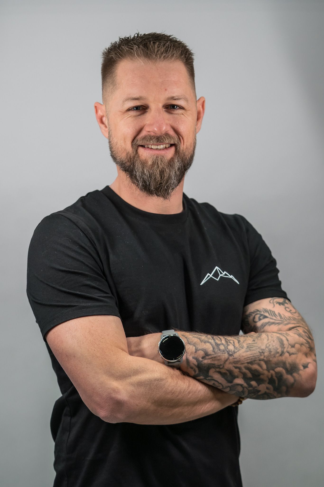 Lächelnder Mann mit tätowierten Armen in einem schwarzen T-Shirt, die Arme verschränkt, vor einem grauen Hintergrund