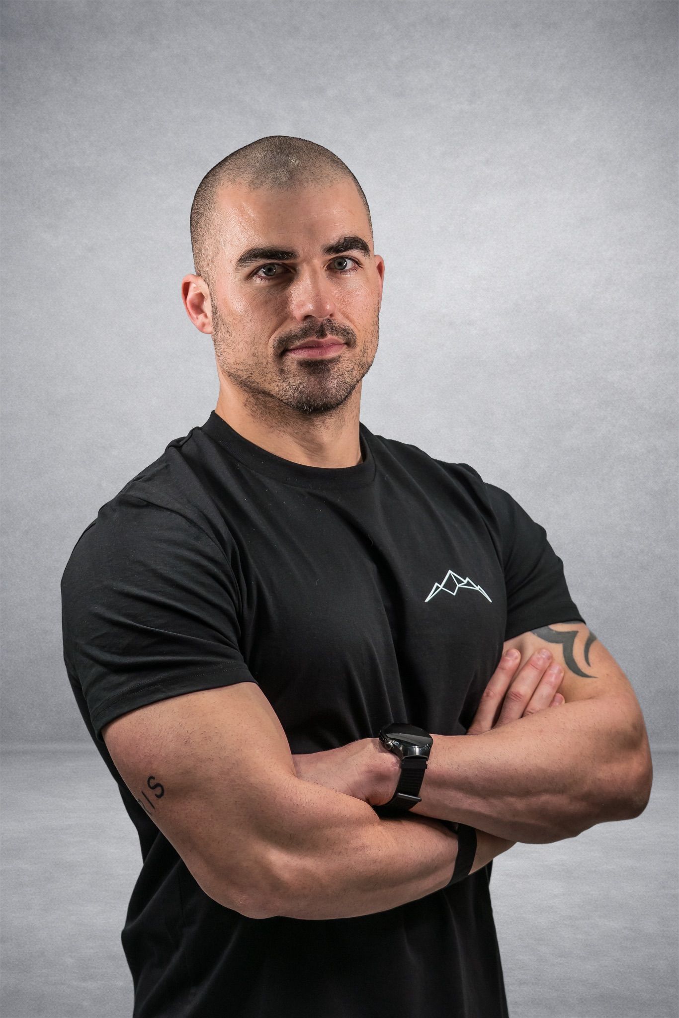 Ein Mann mit verschränkten Armen und einem schwarzen T-Shirt steht vor einem grauen Hintergrund.