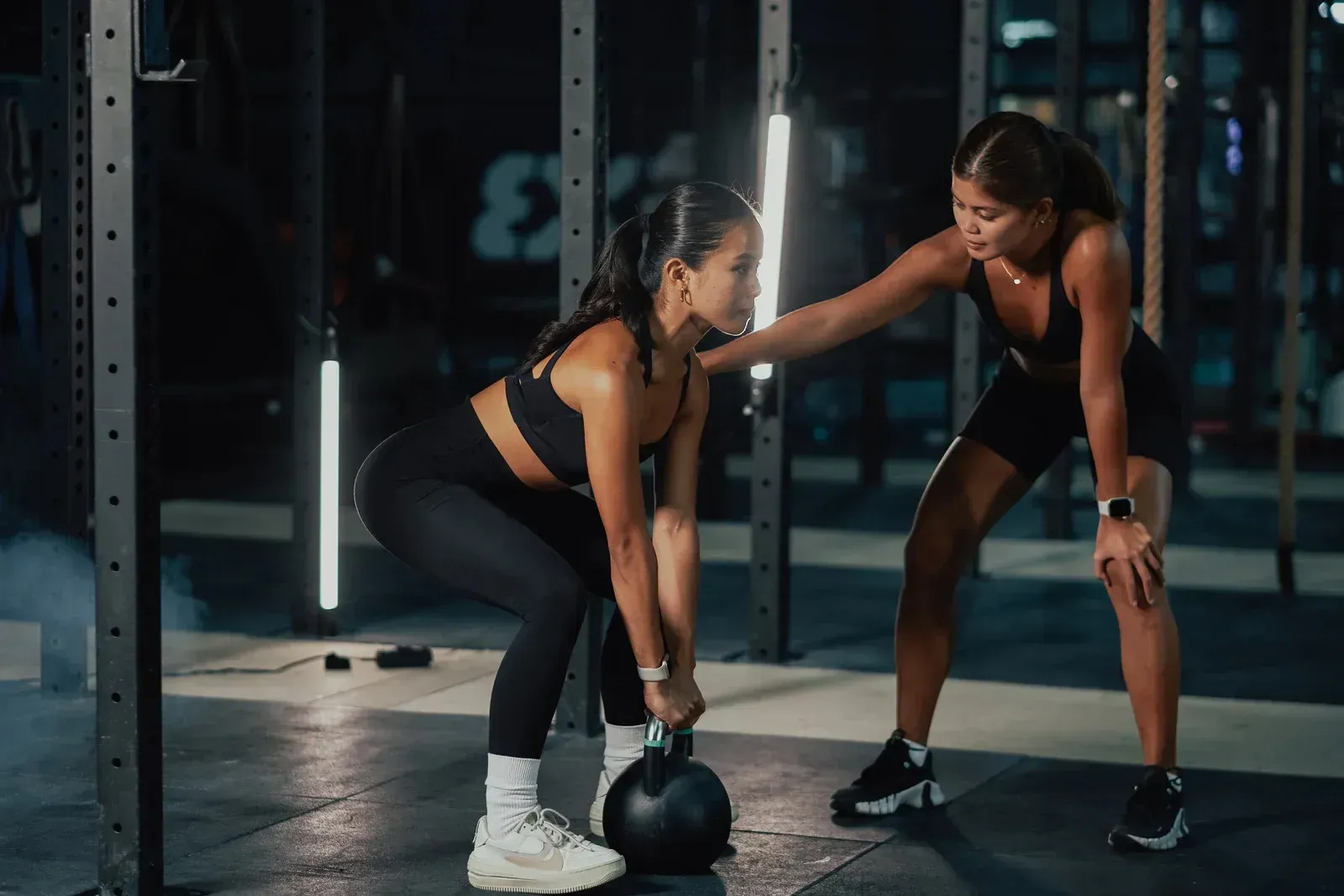 Zwei Frauen in schwarzer Sportkleidung trainieren mit Kettlebells in einem Fitnessstudio.