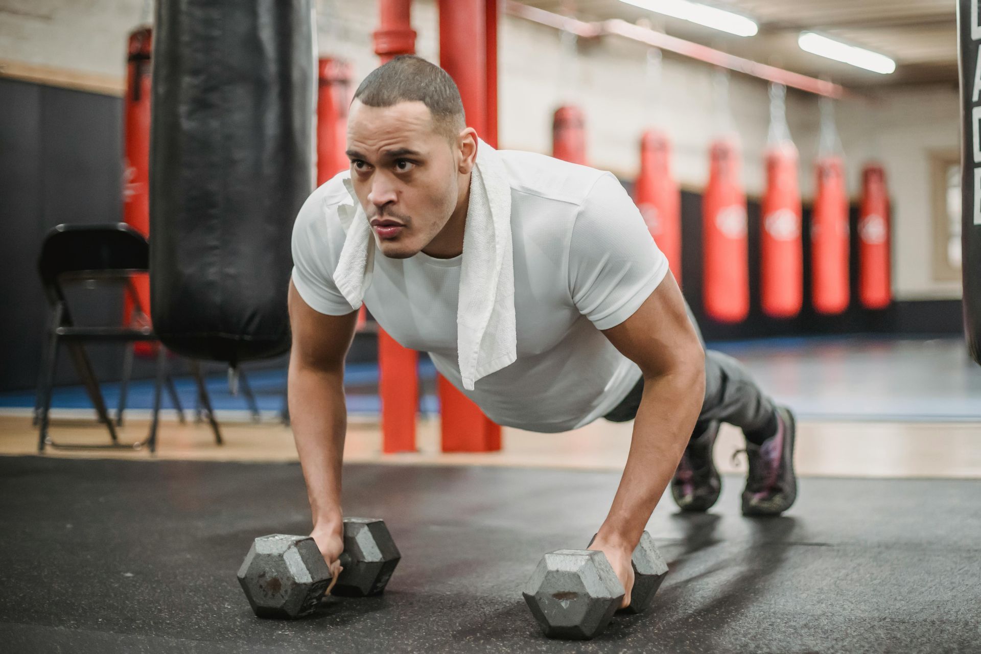 Ein Mann macht Liegestütze mit Kurzhanteln in einem Fitnessstudio; im Hintergrund sind Boxsäcke zu sehen.