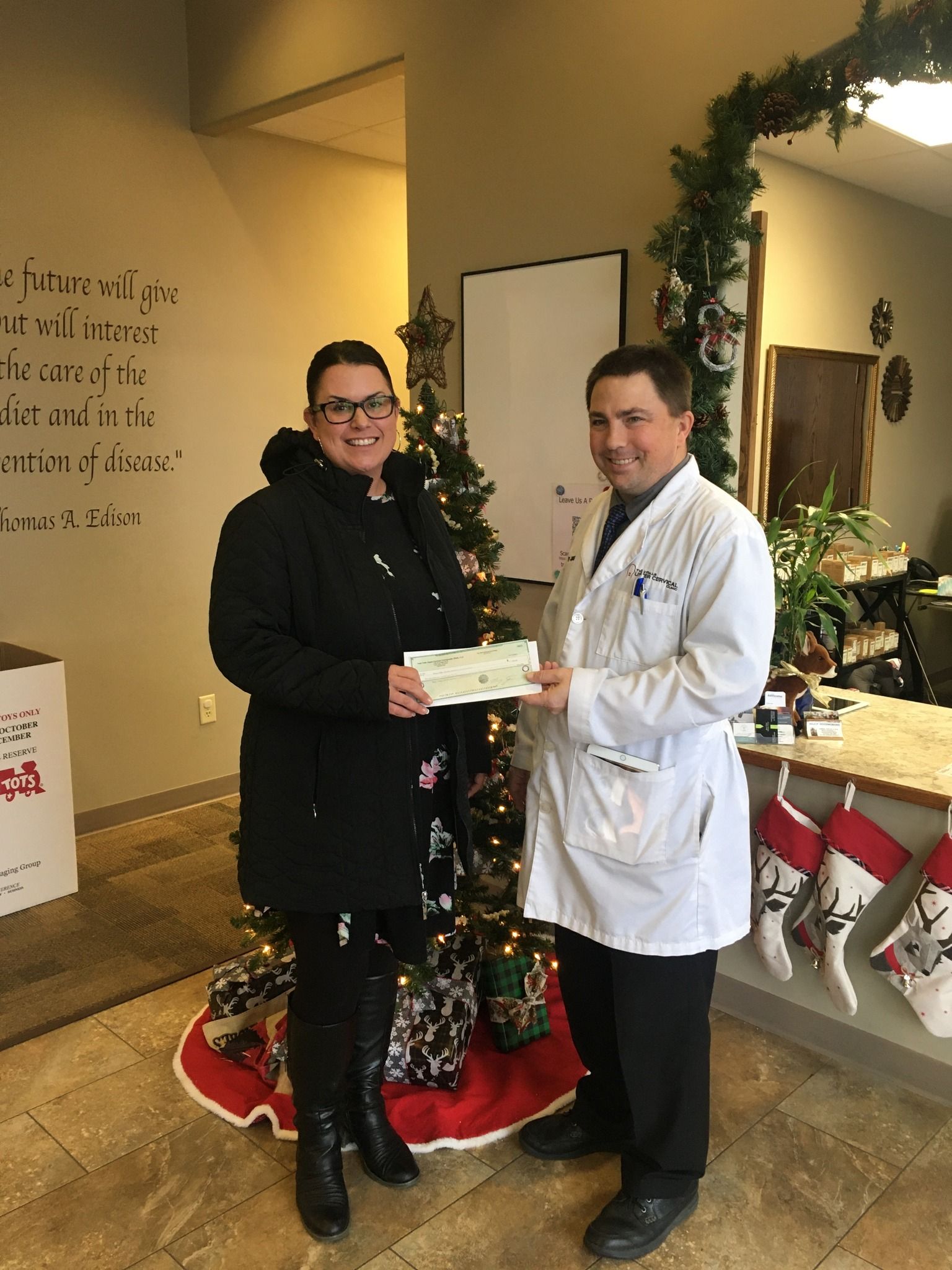 A man in a lab coat is giving a woman a check in front of a christmas tree.