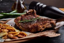 Gegrilltes Tomahawk-Steak mit Pommes frites auf einem Holzbrett, dazu Olivenöl, Paprika und Pfeffermühle.