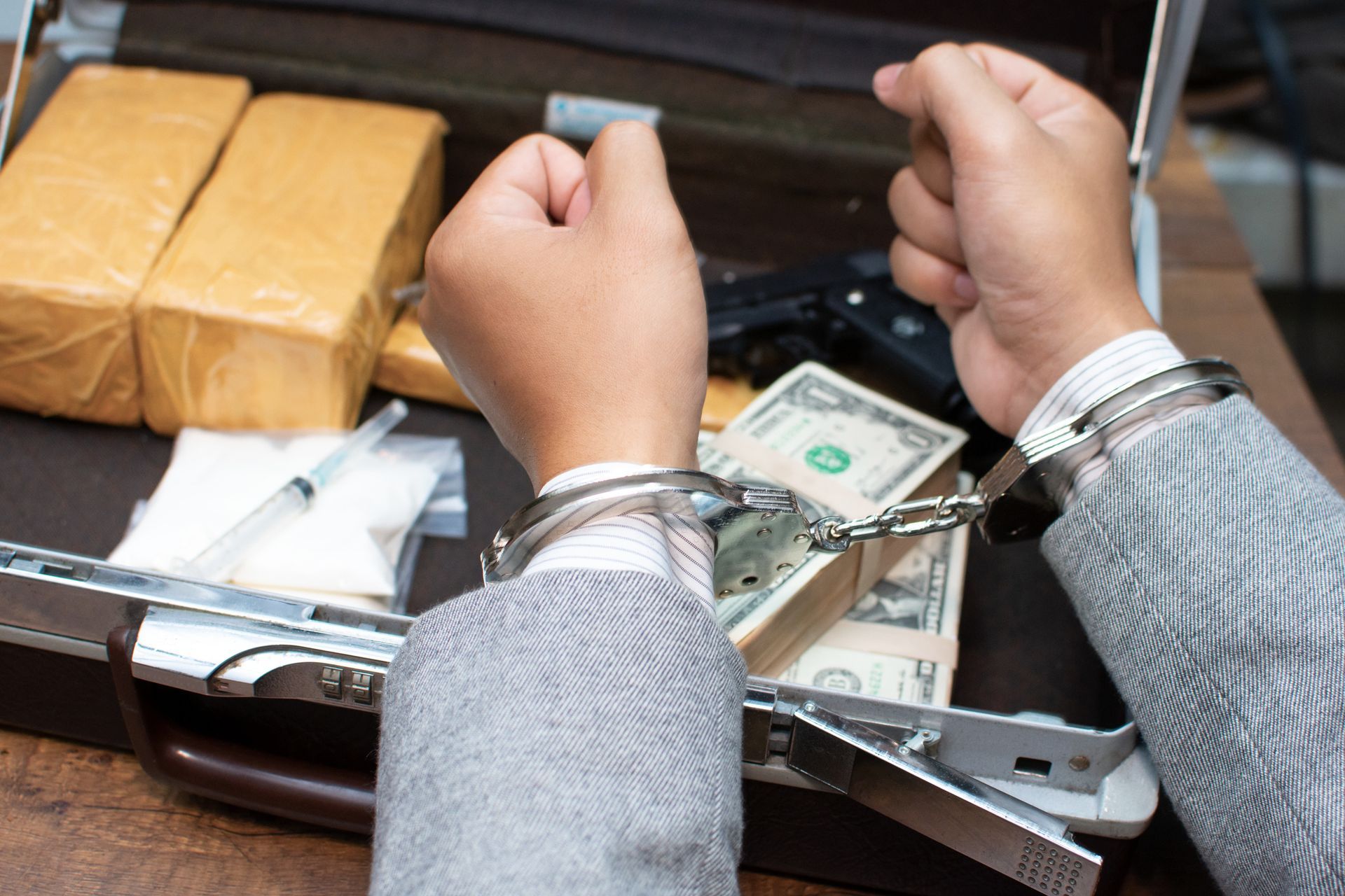 Hands in handcuffs over an open briefcase with drugs, money, and a gun.