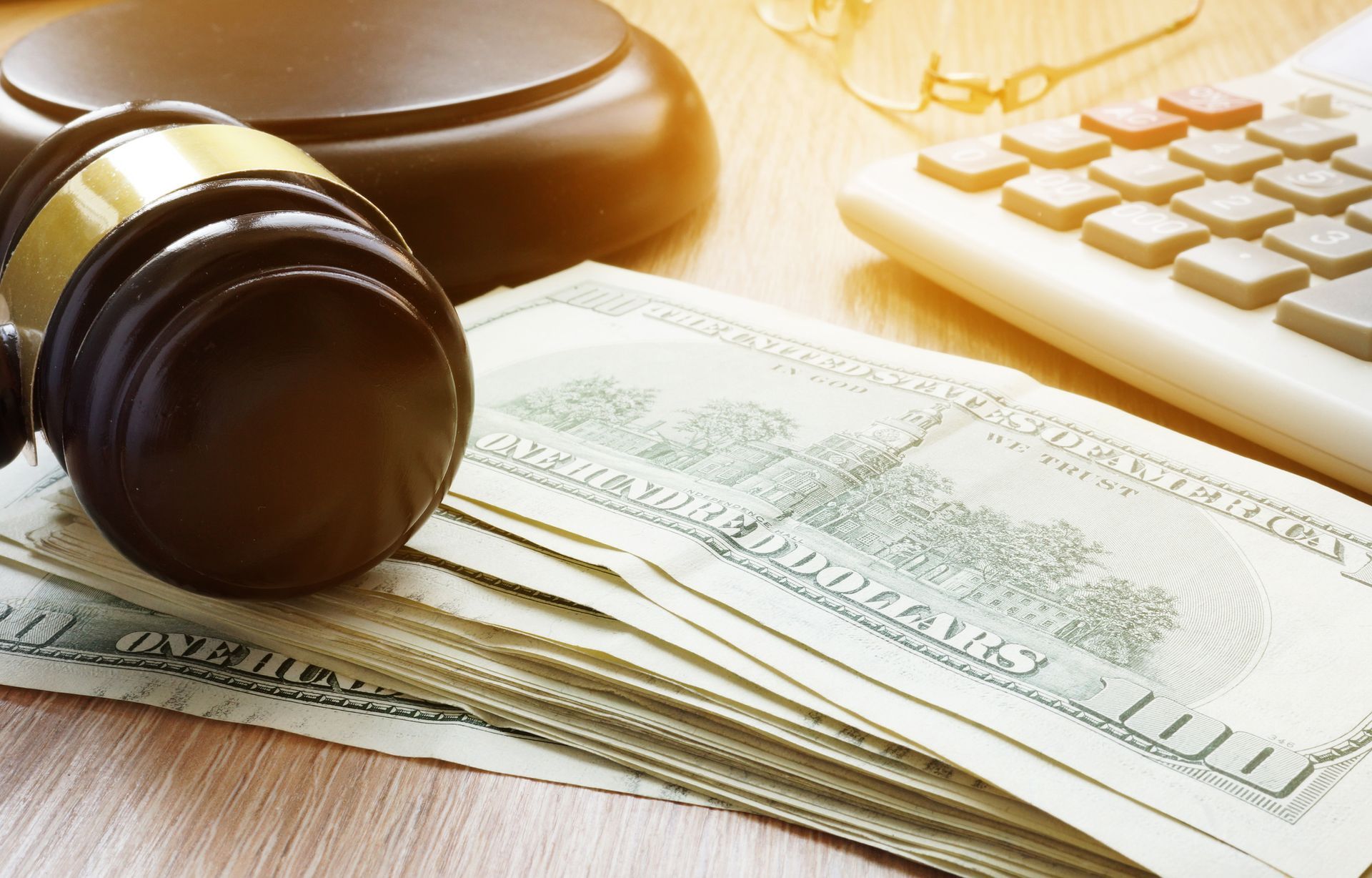 Gavel rests on a stack of cash, next to a calculator and eyeglasses, on a wooden desk.