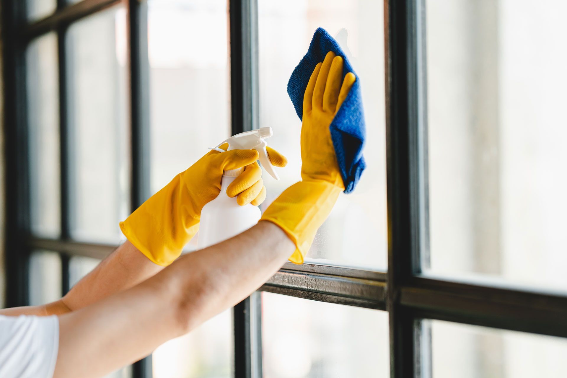 A person wearing yellow gloves is cleaning a window with a spray bottle and a cloth.