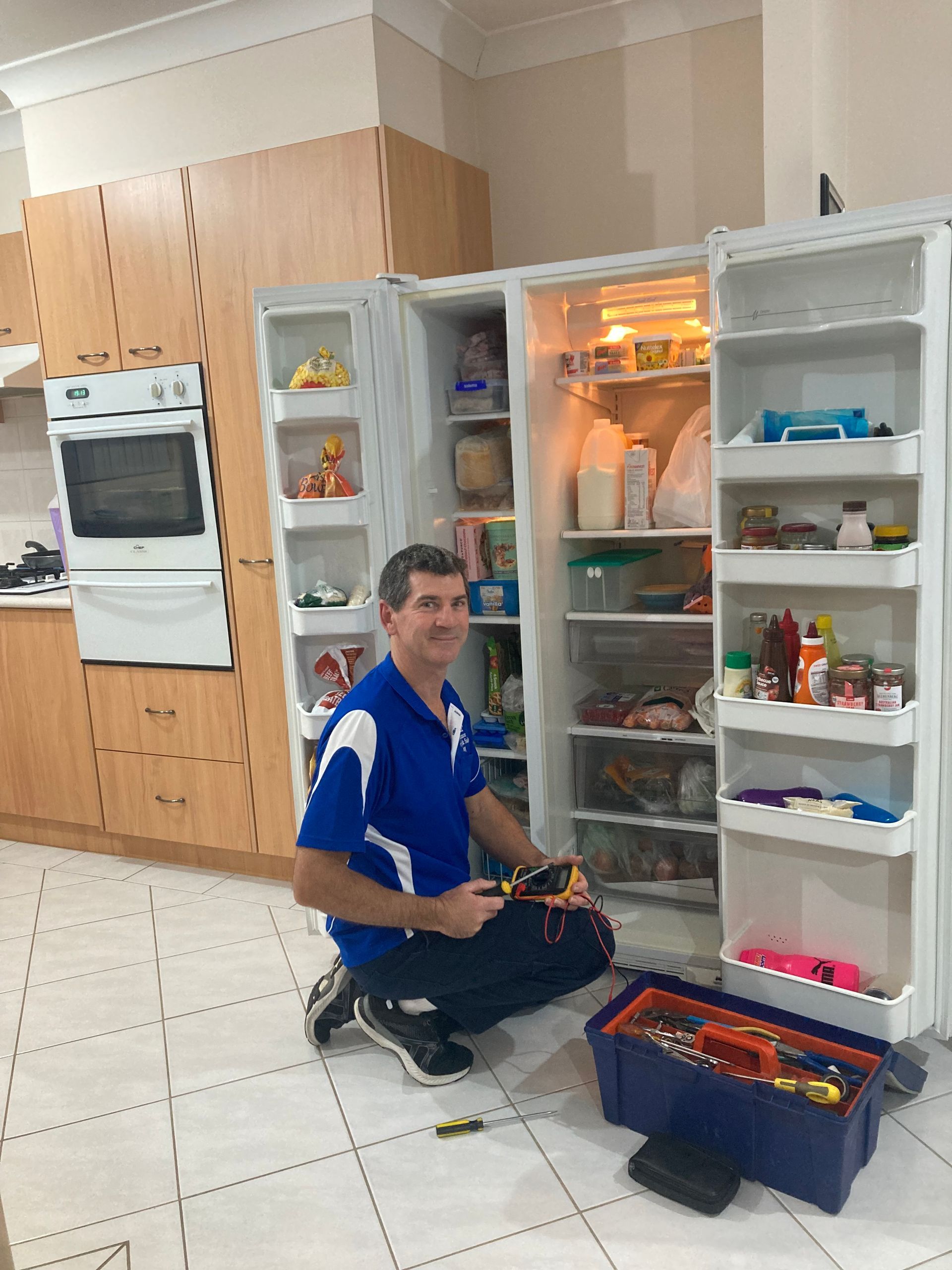 A repairman kneels, fixing a refrigerator with open doors. He holds tools in a toolbox in a kitchen — A.S.A.P. Appliance Service & Sales in Hamlyn Terrace, NSW