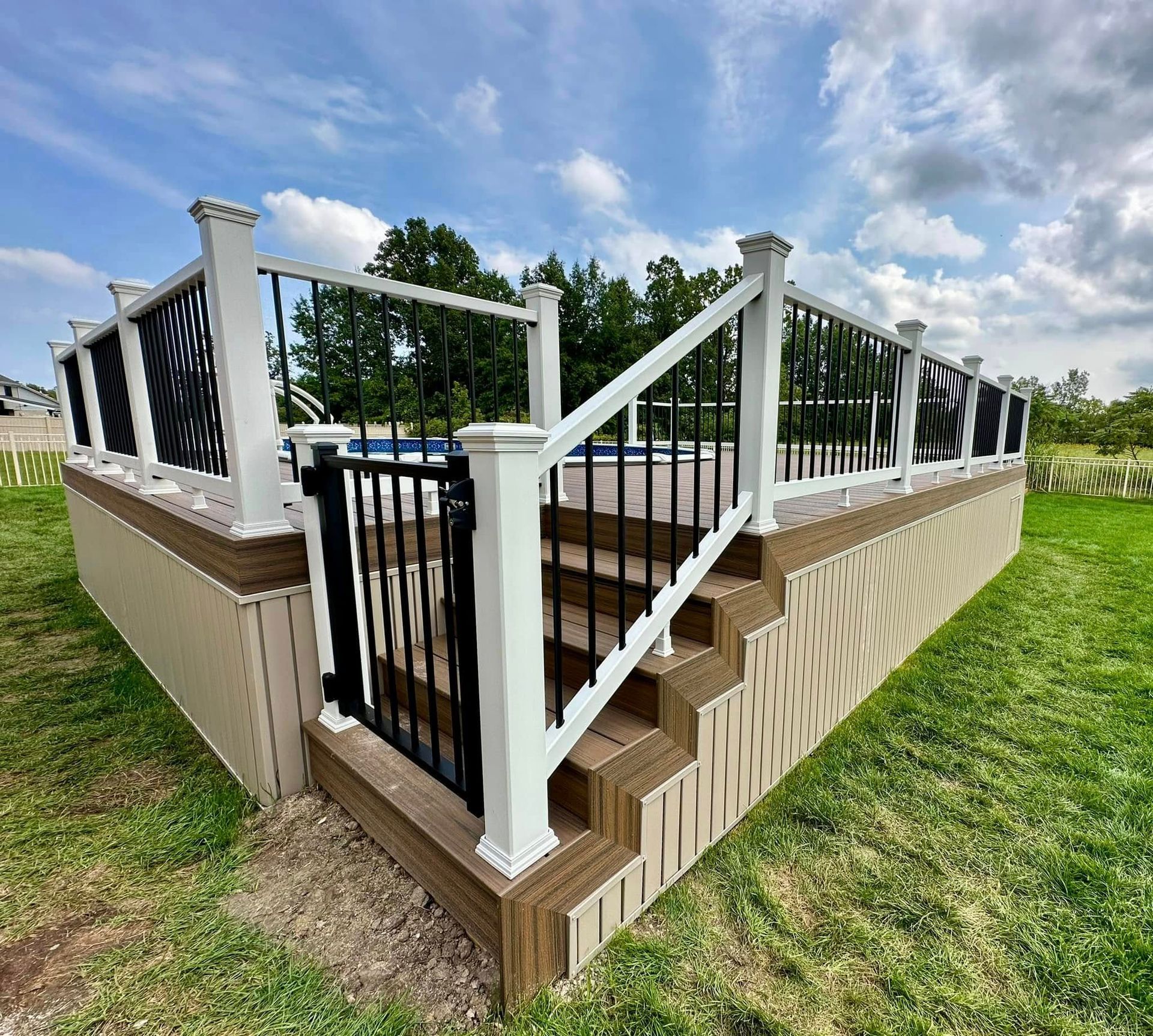Above-ground pool deck with steps and black railing, surrounded by grass under a cloudy sky.