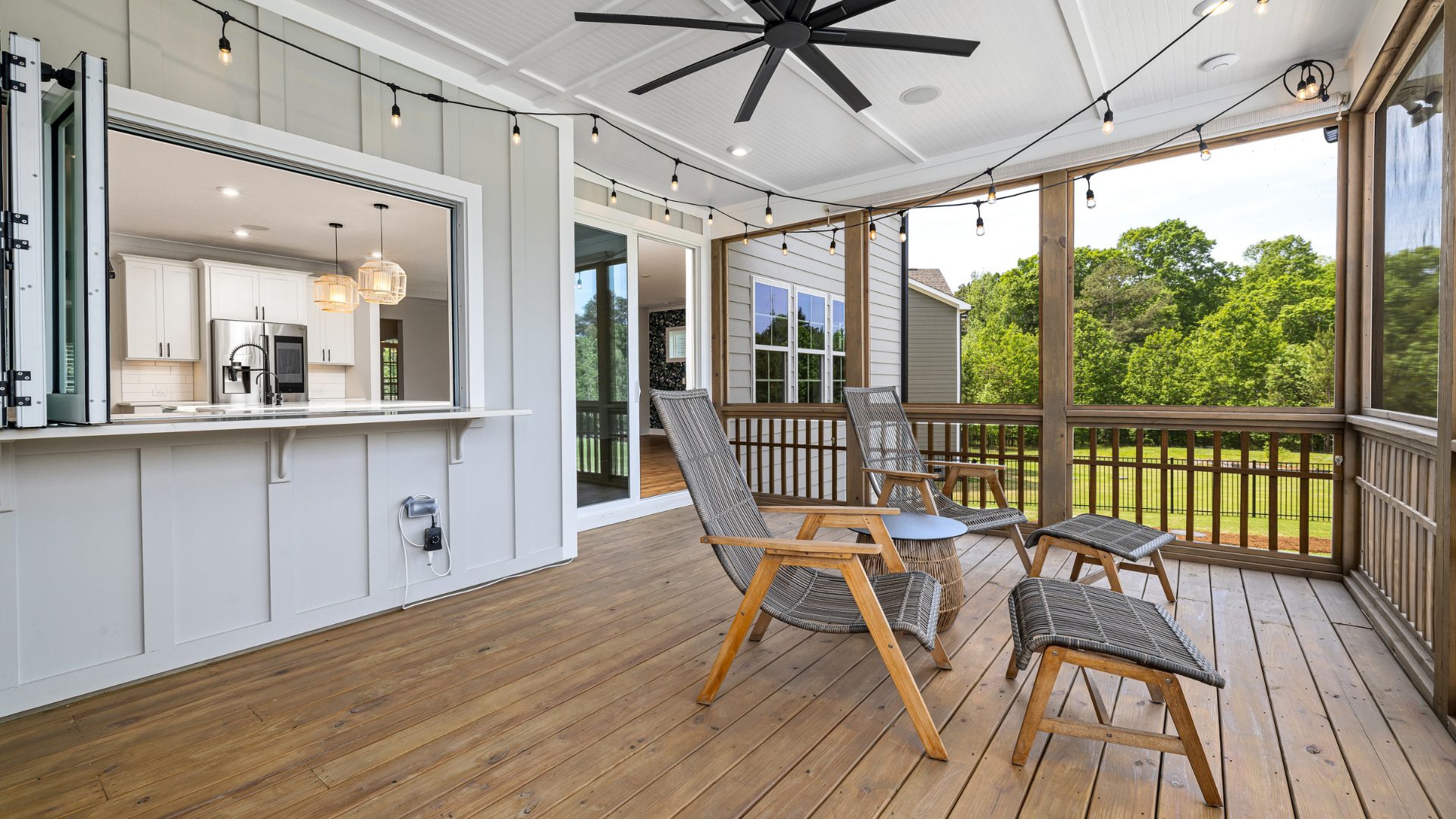 Screened-in porch with wood flooring, open to kitchen. Lounge chairs and string lights visible.