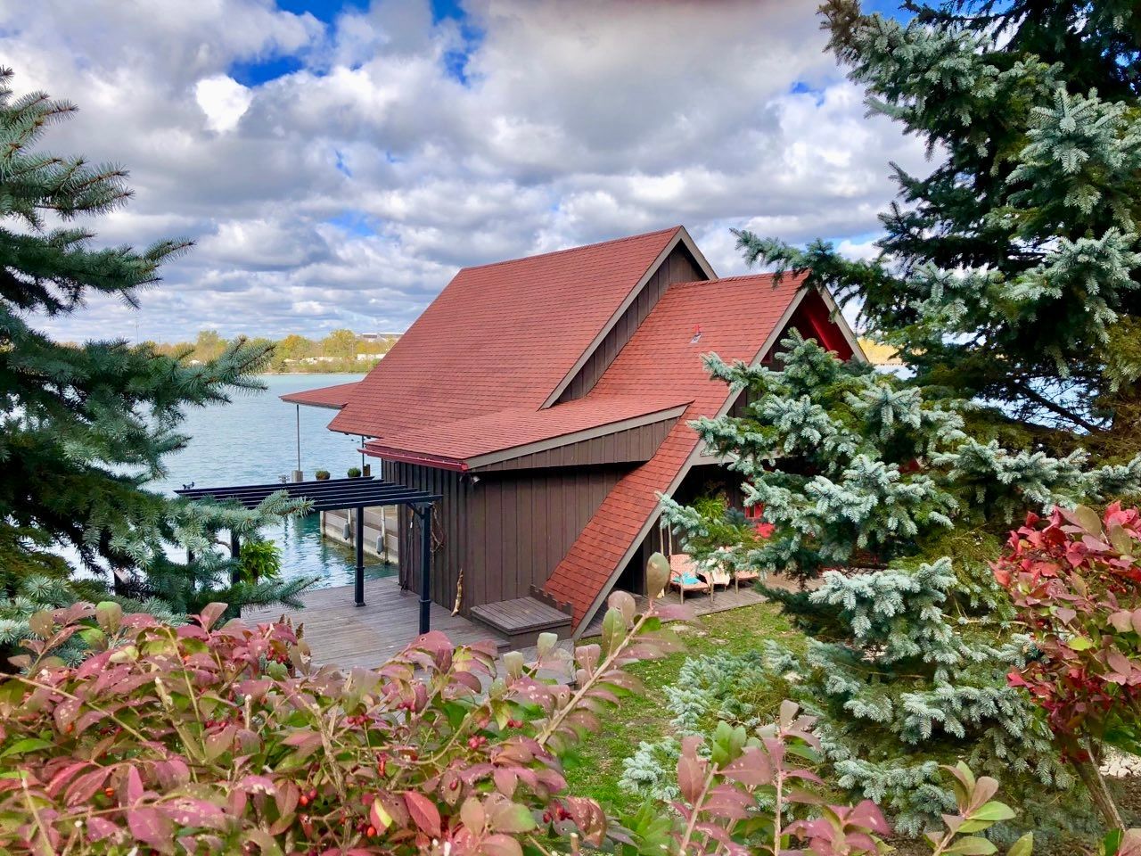 Brown cabin with red roof by water, framed by trees and bushes. Overcast sky.