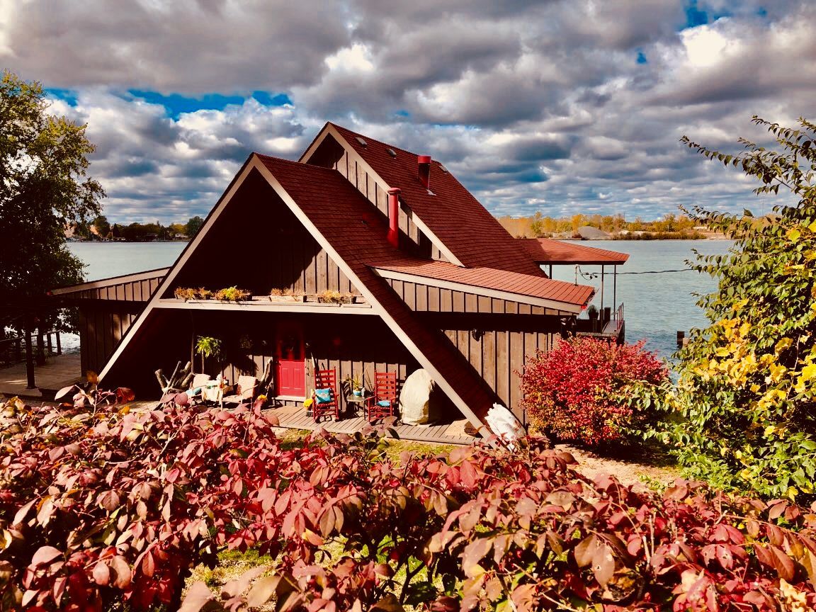 A-frame cabin by lake with red roof, surrounded by fall foliage under a cloudy sky.