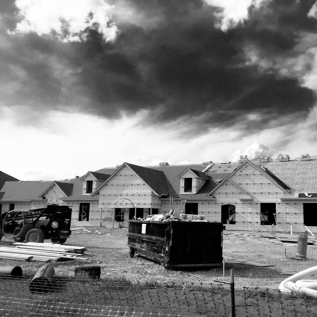 Black and white photo of new homes under construction with a dumpster and equipment in the foreground, cloudy sky.