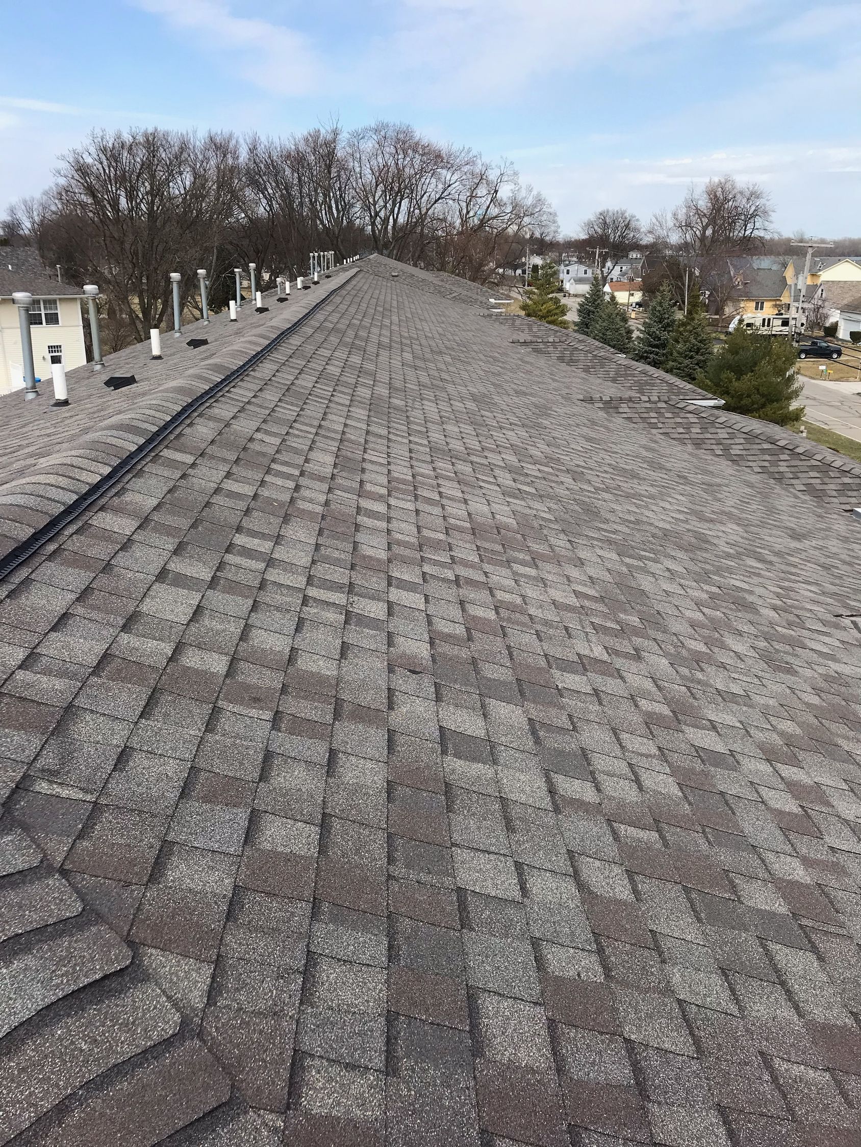 View of a weathered asphalt shingle roof with several vents, trees, and buildings in the background.