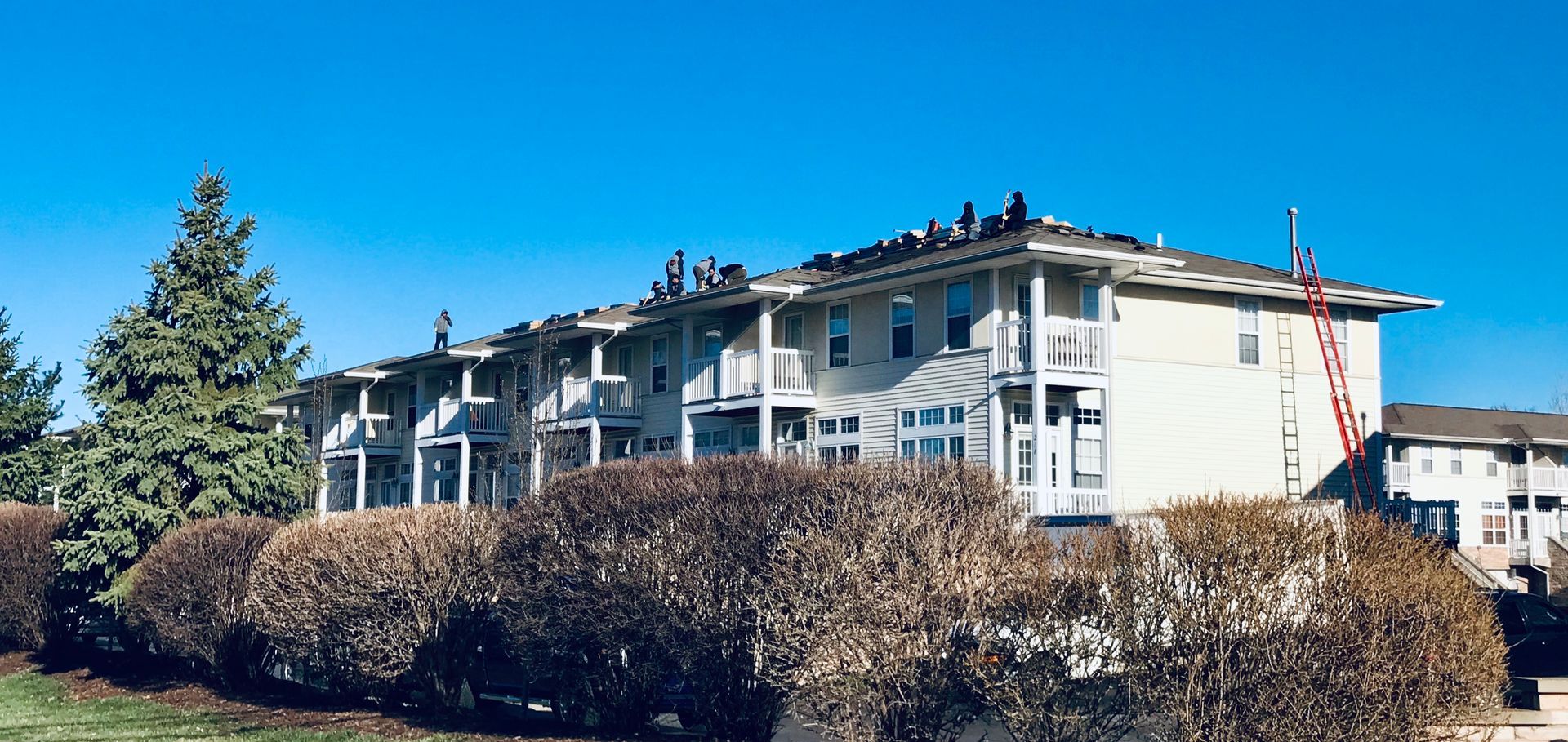 Workers repairing a building roof on a sunny day. A red ladder is leaning against the side.