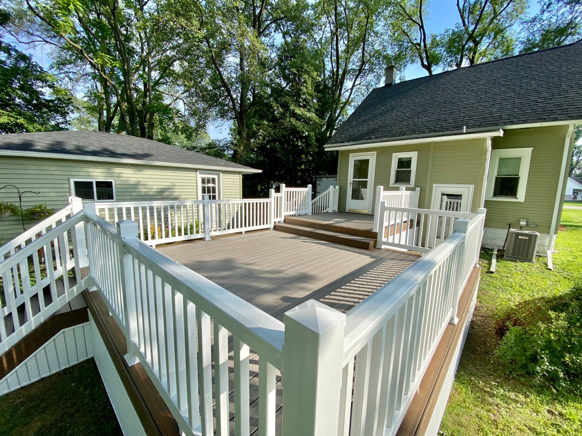 A two-story green house with a white deck. A smaller building is in the background. Sunny day.