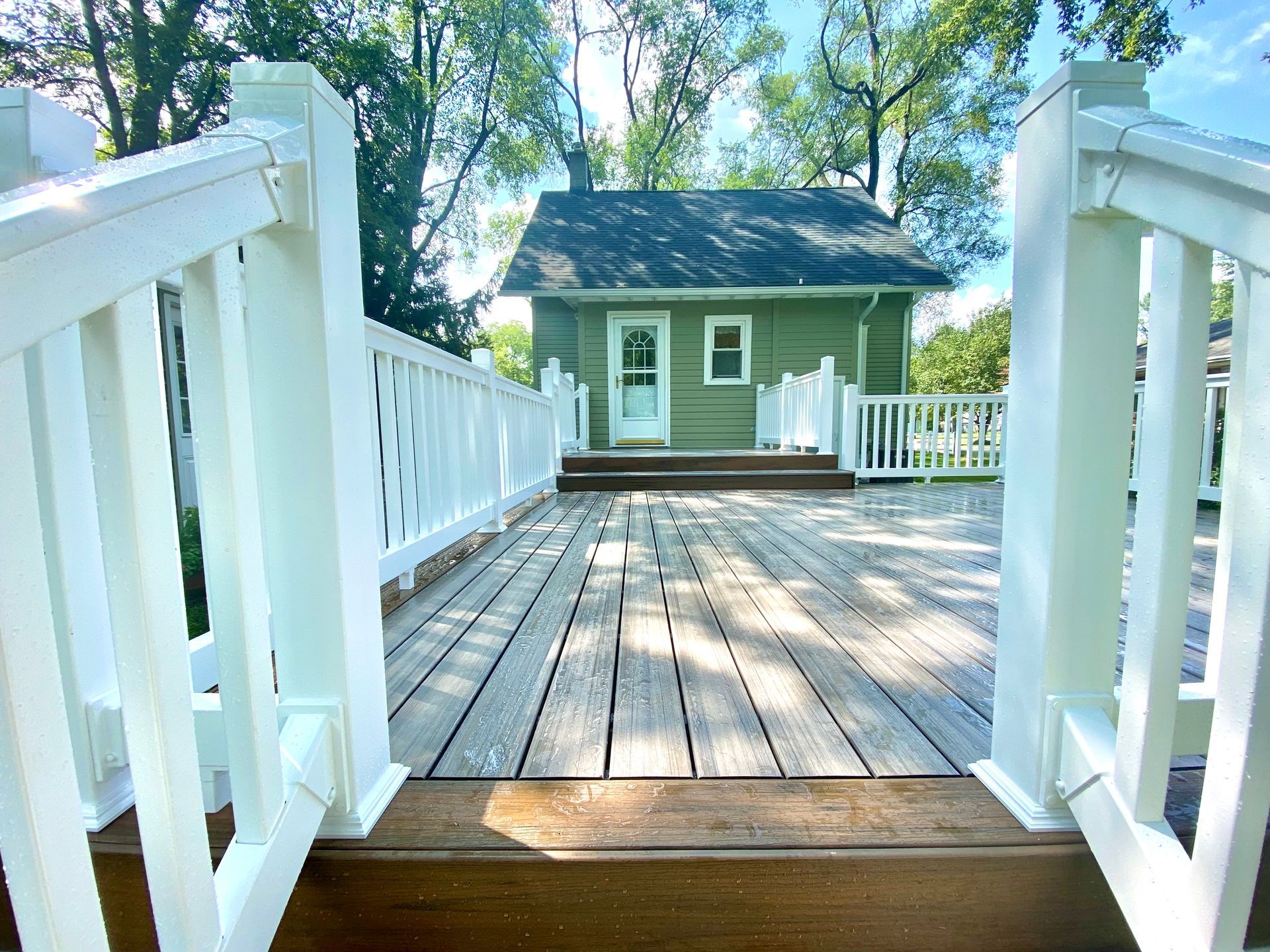 Wooden deck with white railings, leading to a small, green house with a white door.