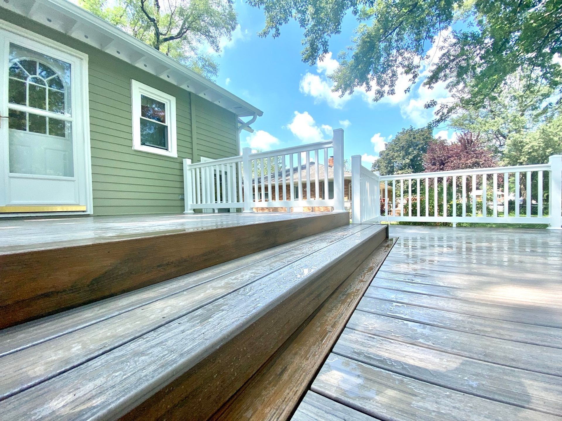 Wooden deck with steps leading to a green house, white railing, and blue sky.