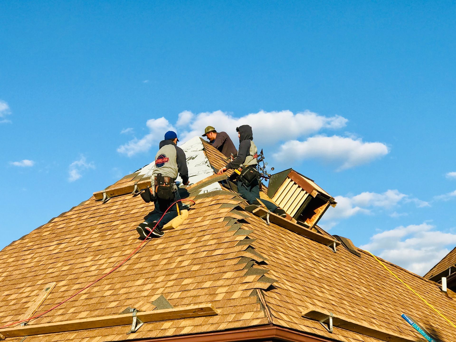 Roofers installing shingles on a house roof under a blue sky.