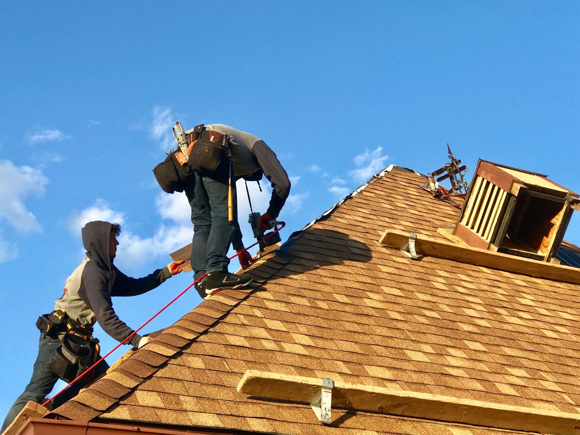 Two roofers working on a brown shingle roof under a blue sky, one secured with a safety harness.