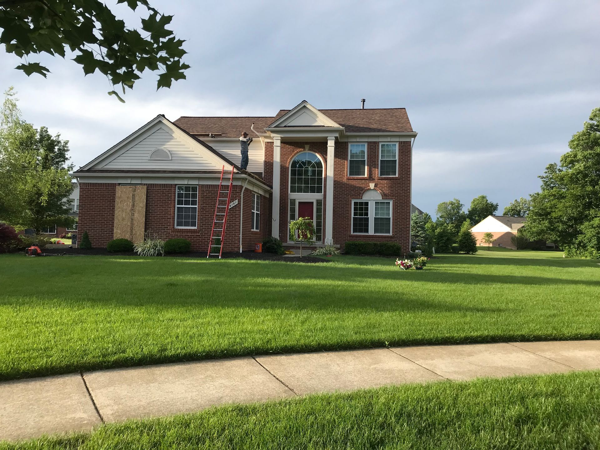 Two-story red brick house with boarded-up window, green lawn, and cloudy sky.
