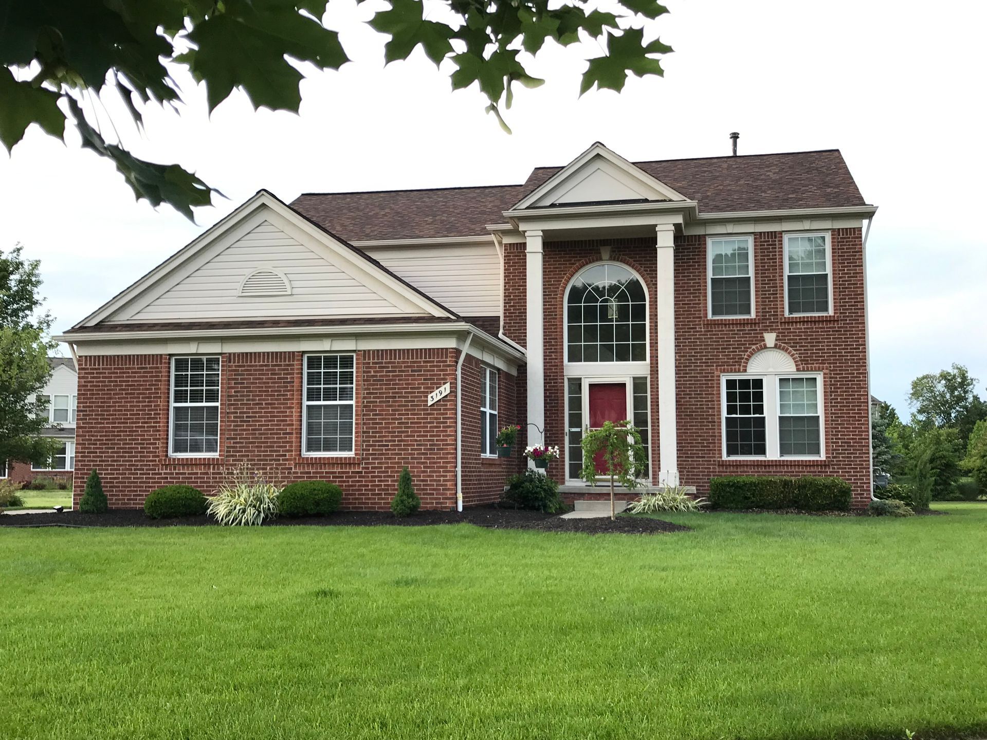 Two-story brick house with a green lawn and brown roof under a cloudy sky.