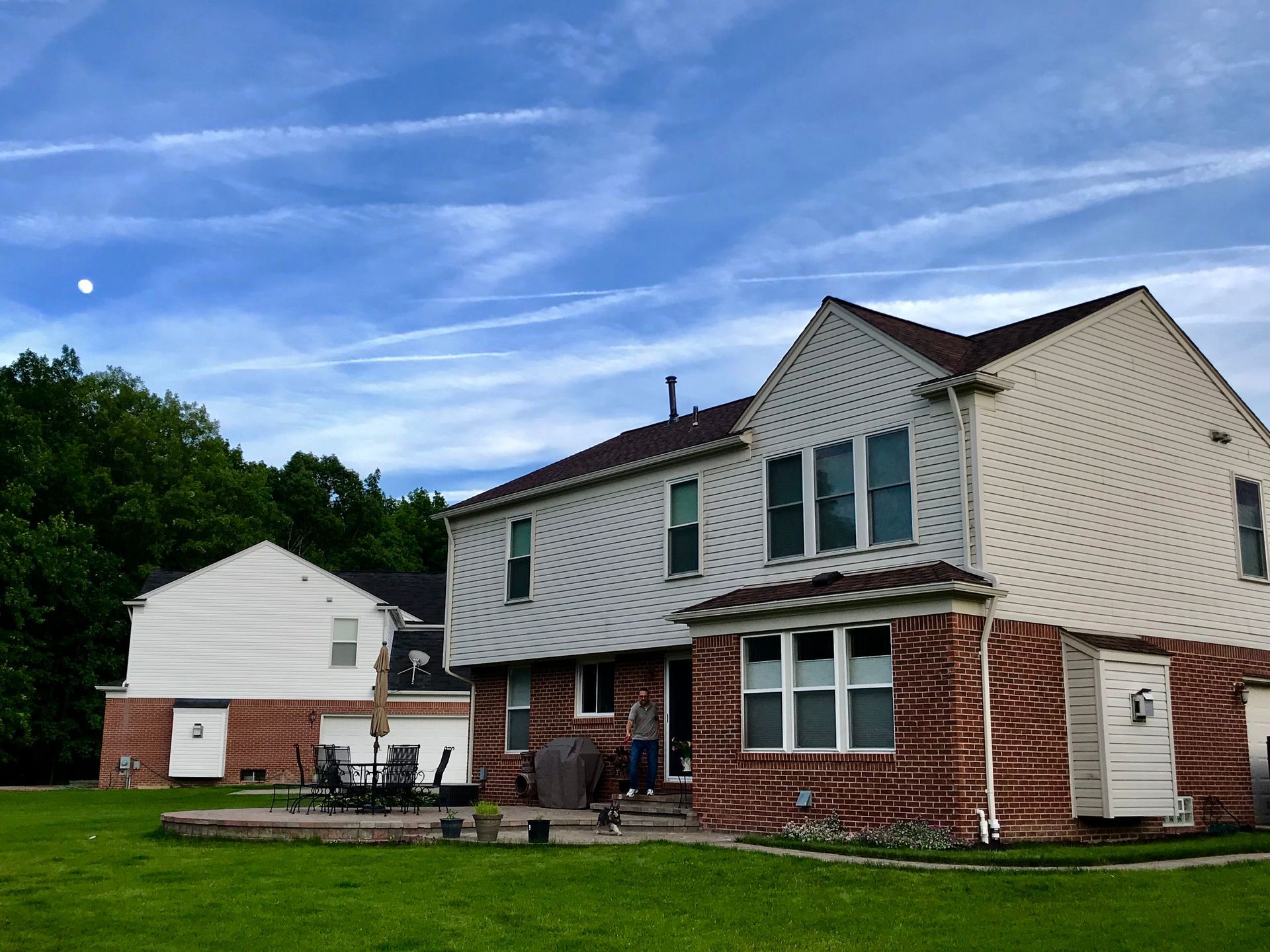 Backyard of a two-story house with green lawn, trees, patio, and blue sky.