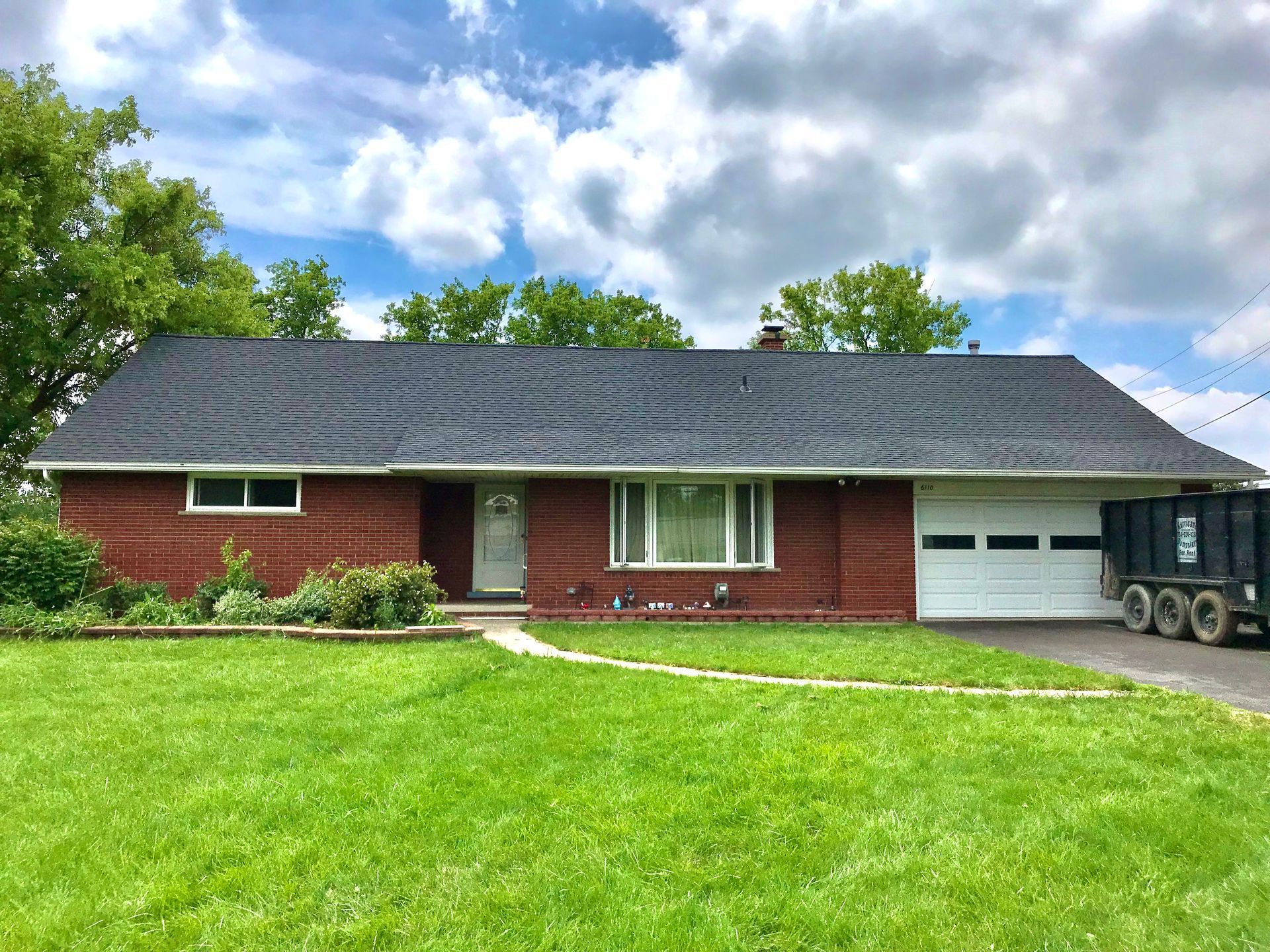 Red brick ranch-style house with black wavy roof, white garage door, and green lawn under a cloudy sky.