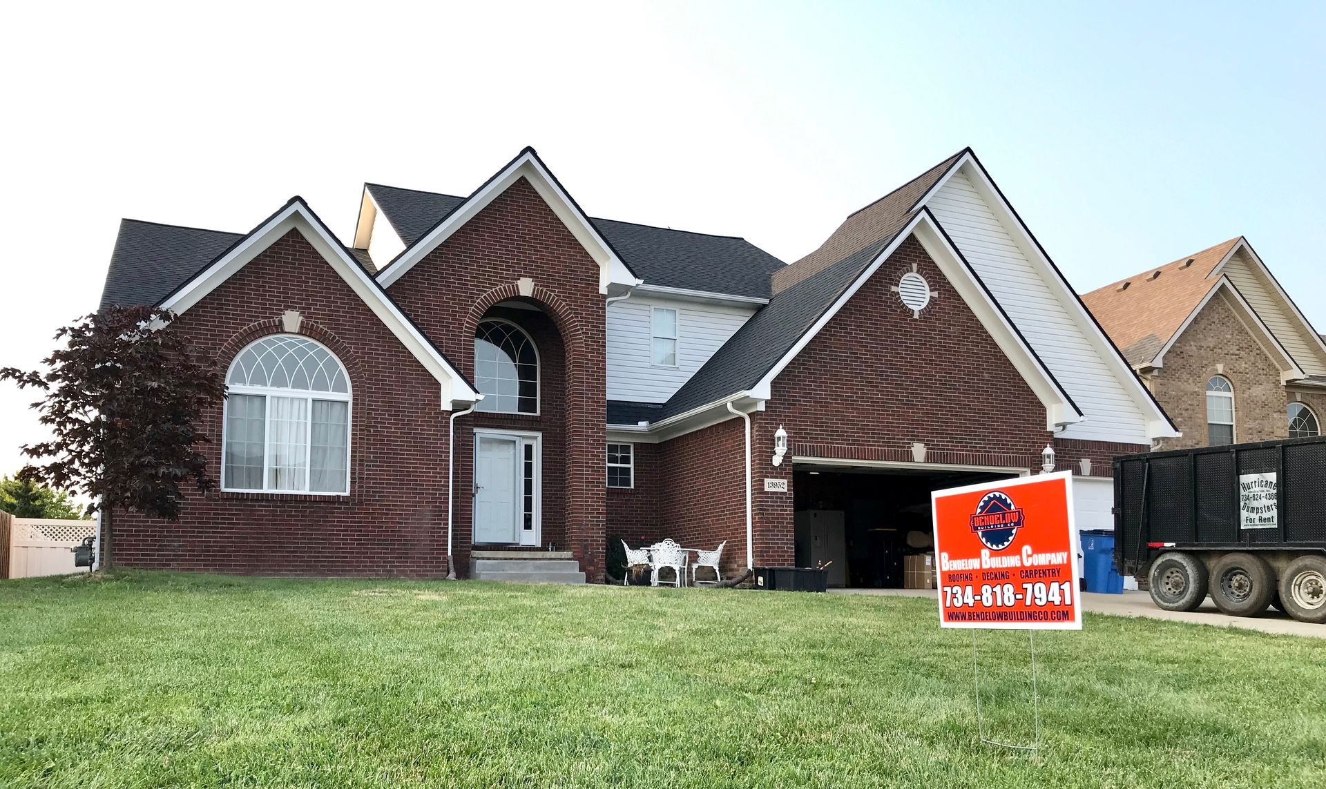 Red brick house with black roof, sign on lawn, and dumpster in driveway.