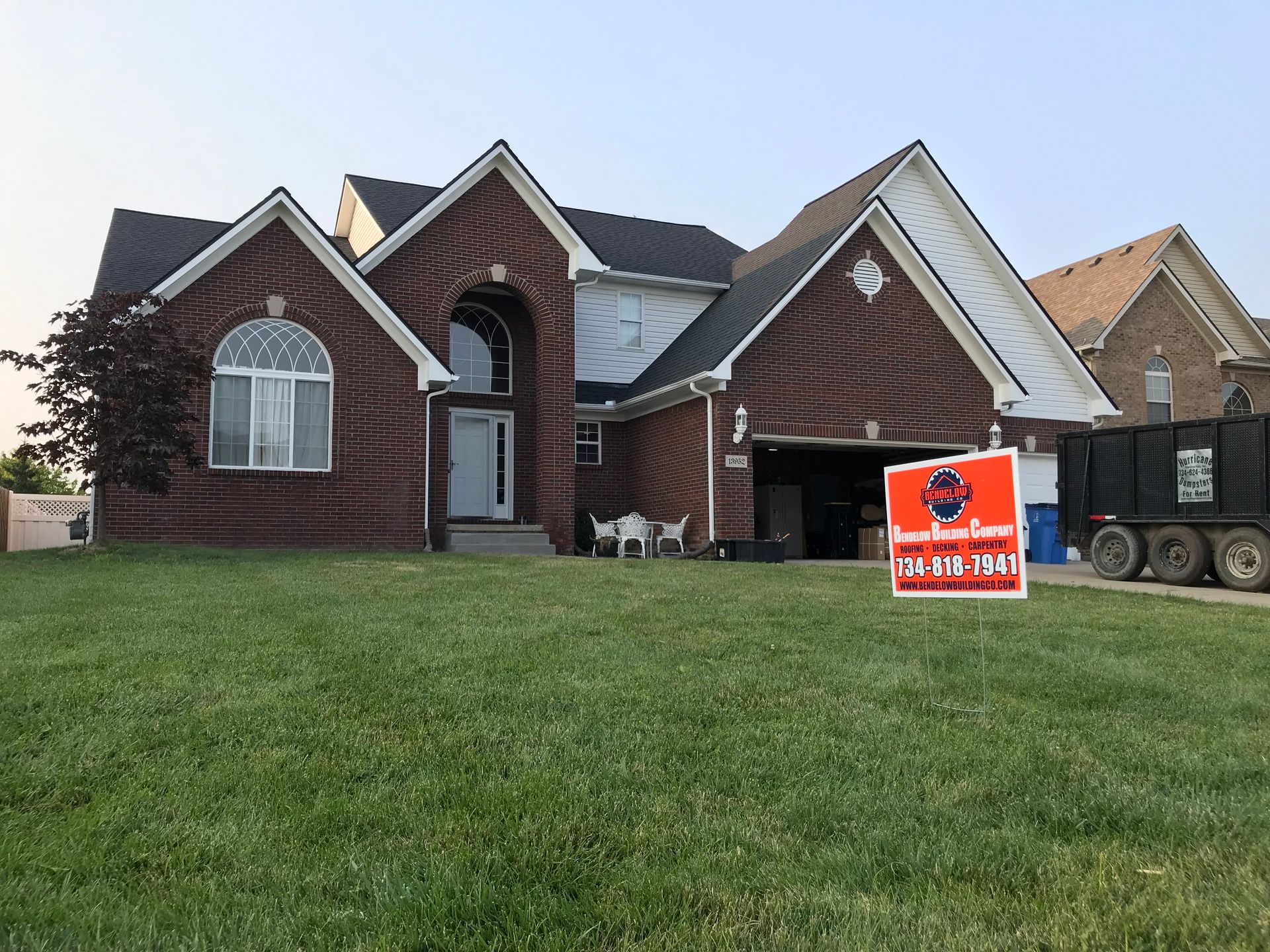 Brick house with open garage door, lawn, and moving services sign. Dumpster visible.