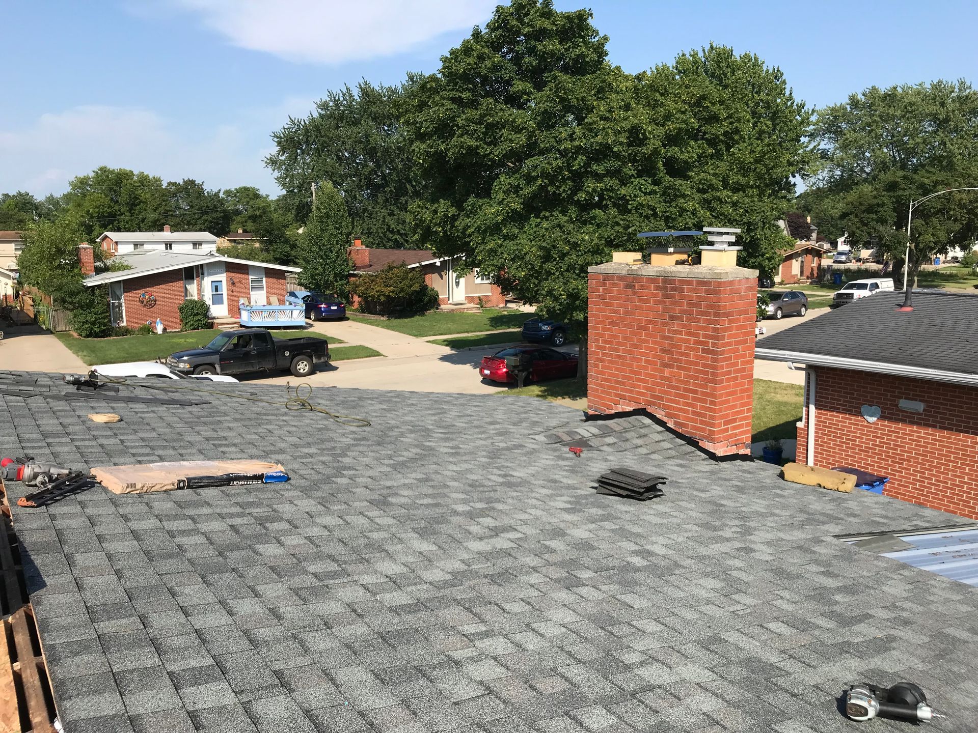 New shingles on a roof with a brick chimney. Other houses and cars are visible in the background on a sunny day.