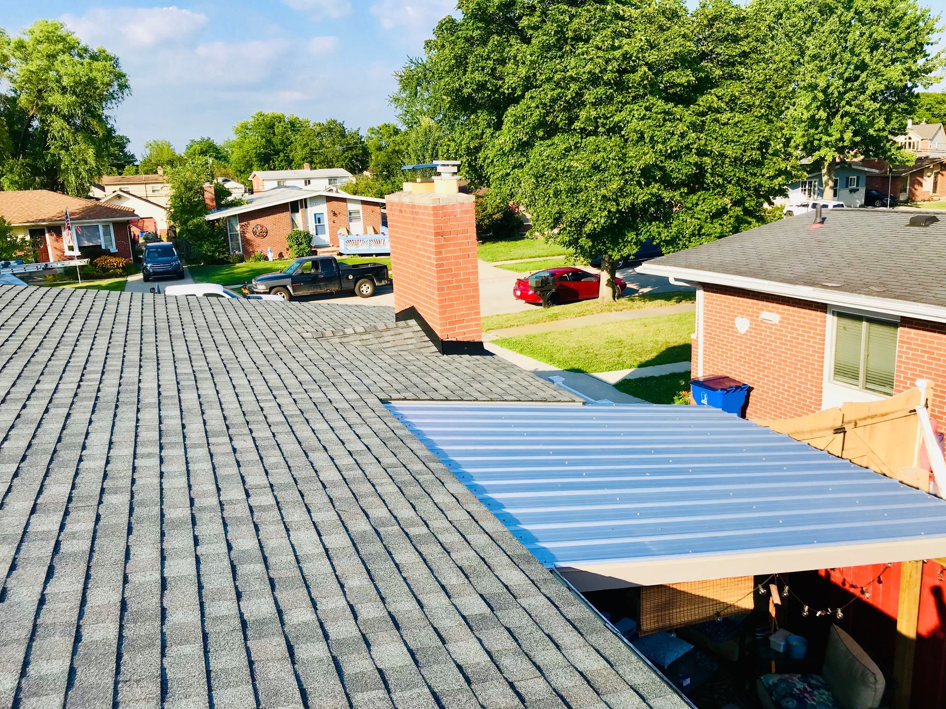 Roof with shingles, chimney, and metal awning on a house in a residential neighborhood with green trees.