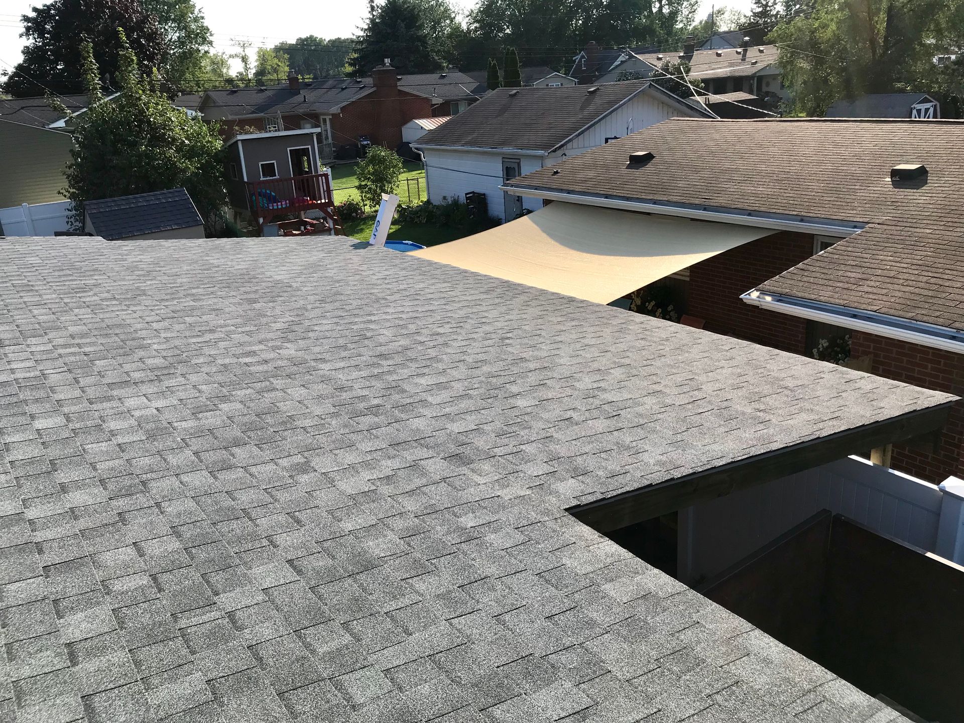 View of a gray shingle roof in a neighborhood, partially obscuring houses and trees under a sunny sky.