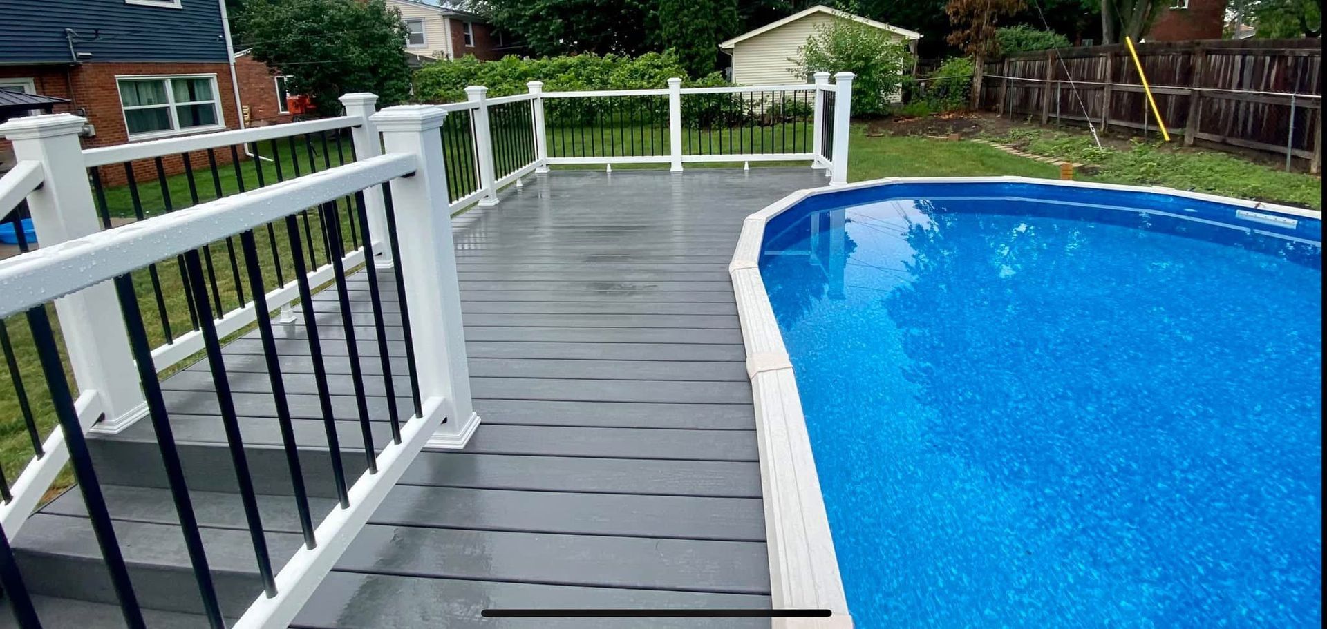 A gray deck next to a blue pool, surrounded by white railings with black spindles.