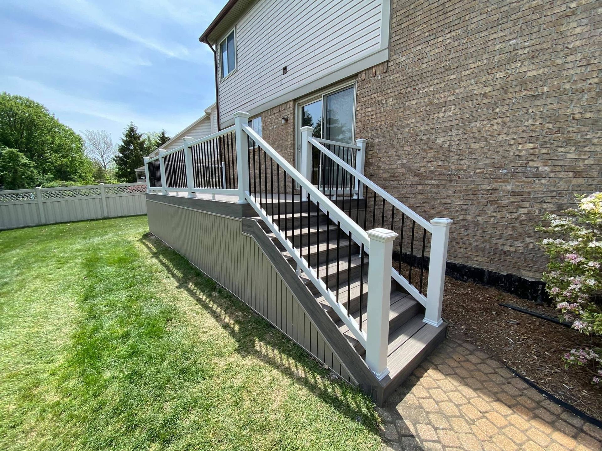 Deck with white and black railings, steps, attached to a brick house, on green grass.