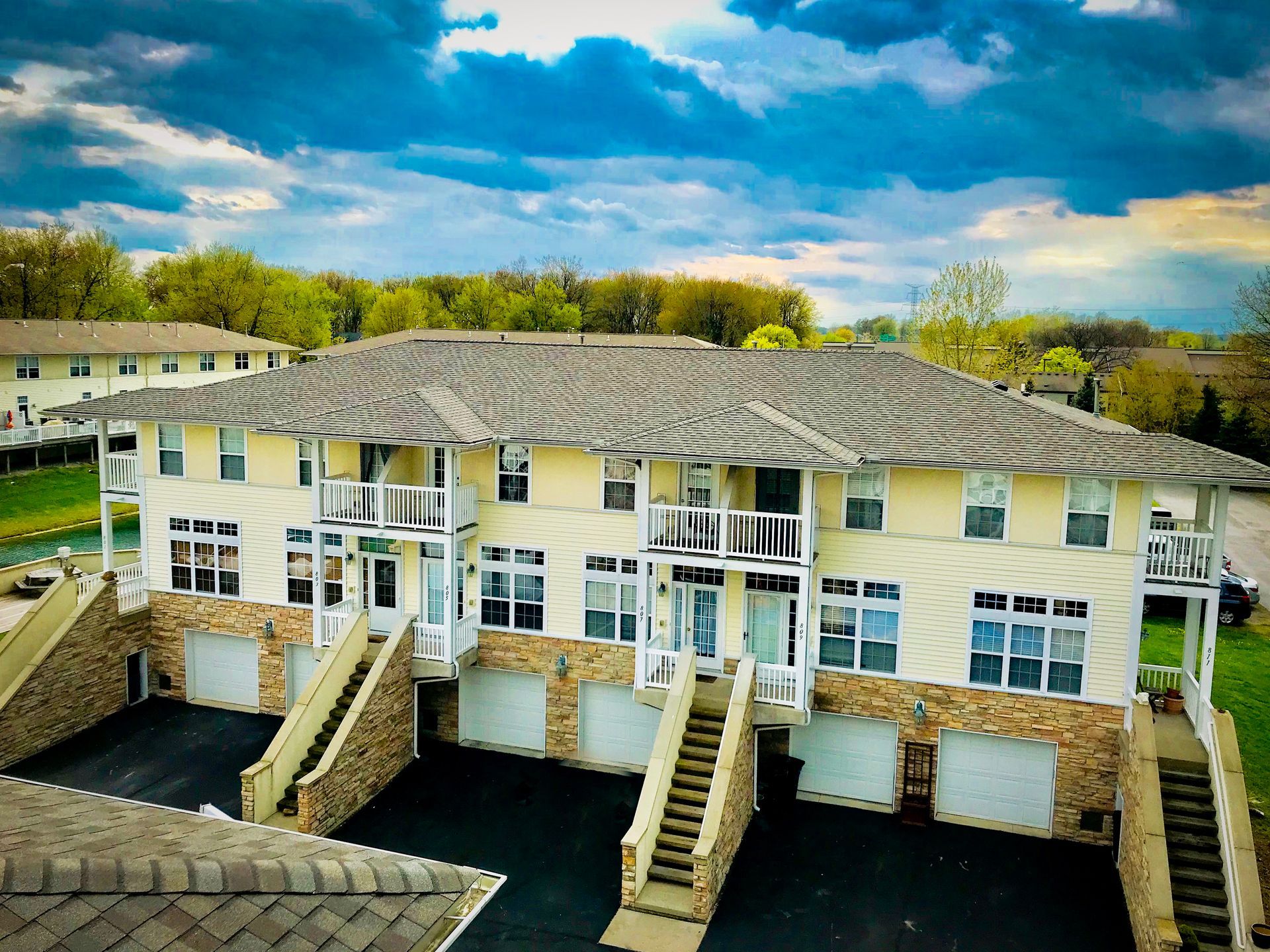 Two-story apartment building with tan siding, stone accents, balconies, and garage doors. Overcast sky in the background.