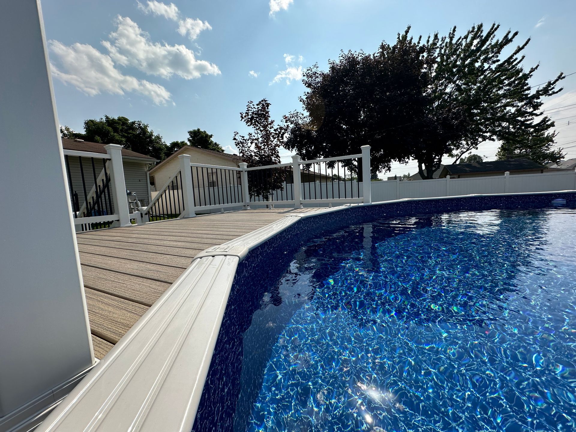 Above-ground swimming pool with blue water and wooden deck on a sunny day. White railing surrounds it.