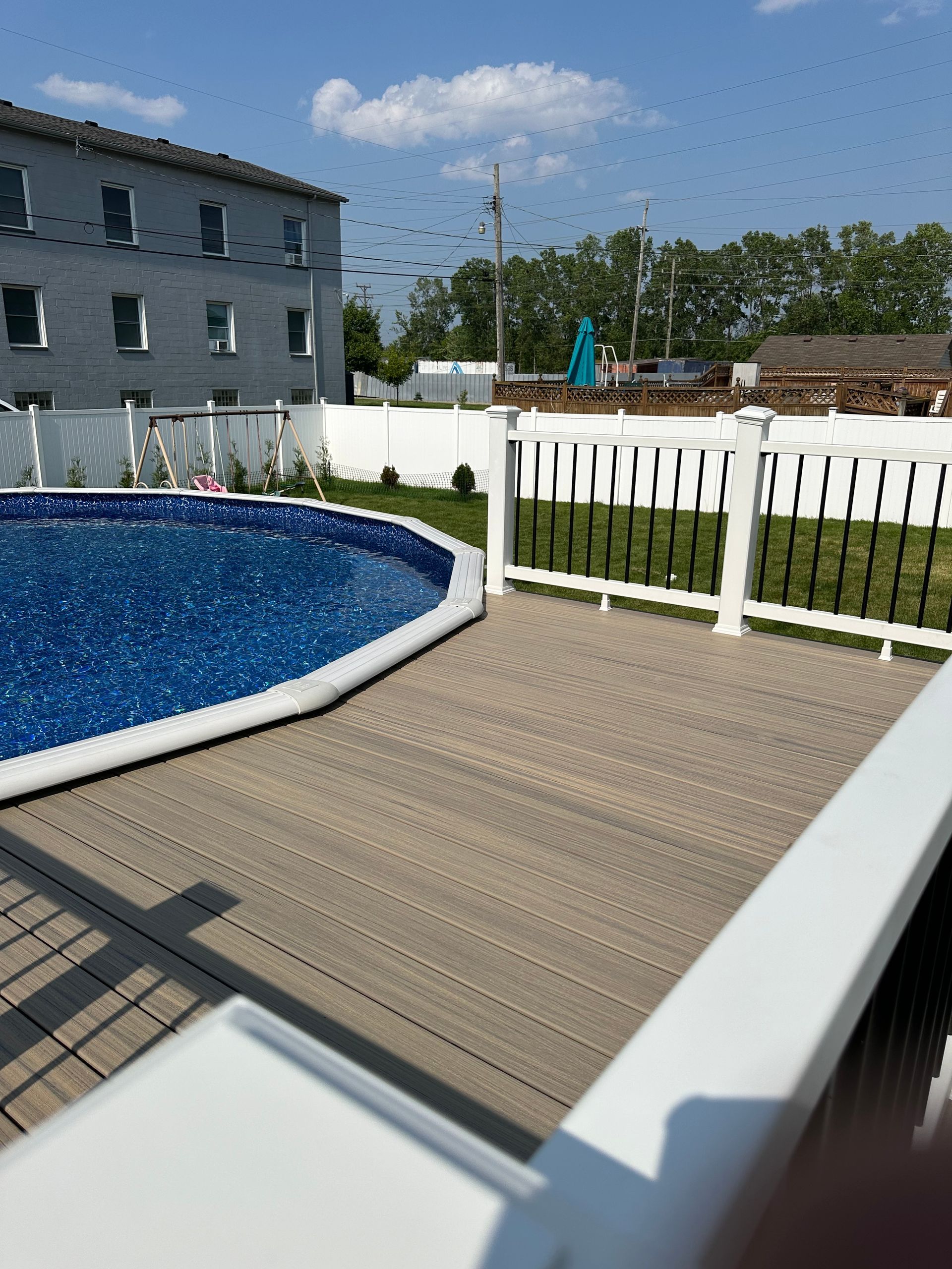 Above-ground pool with a light gray deck and white fence under a partly cloudy sky.