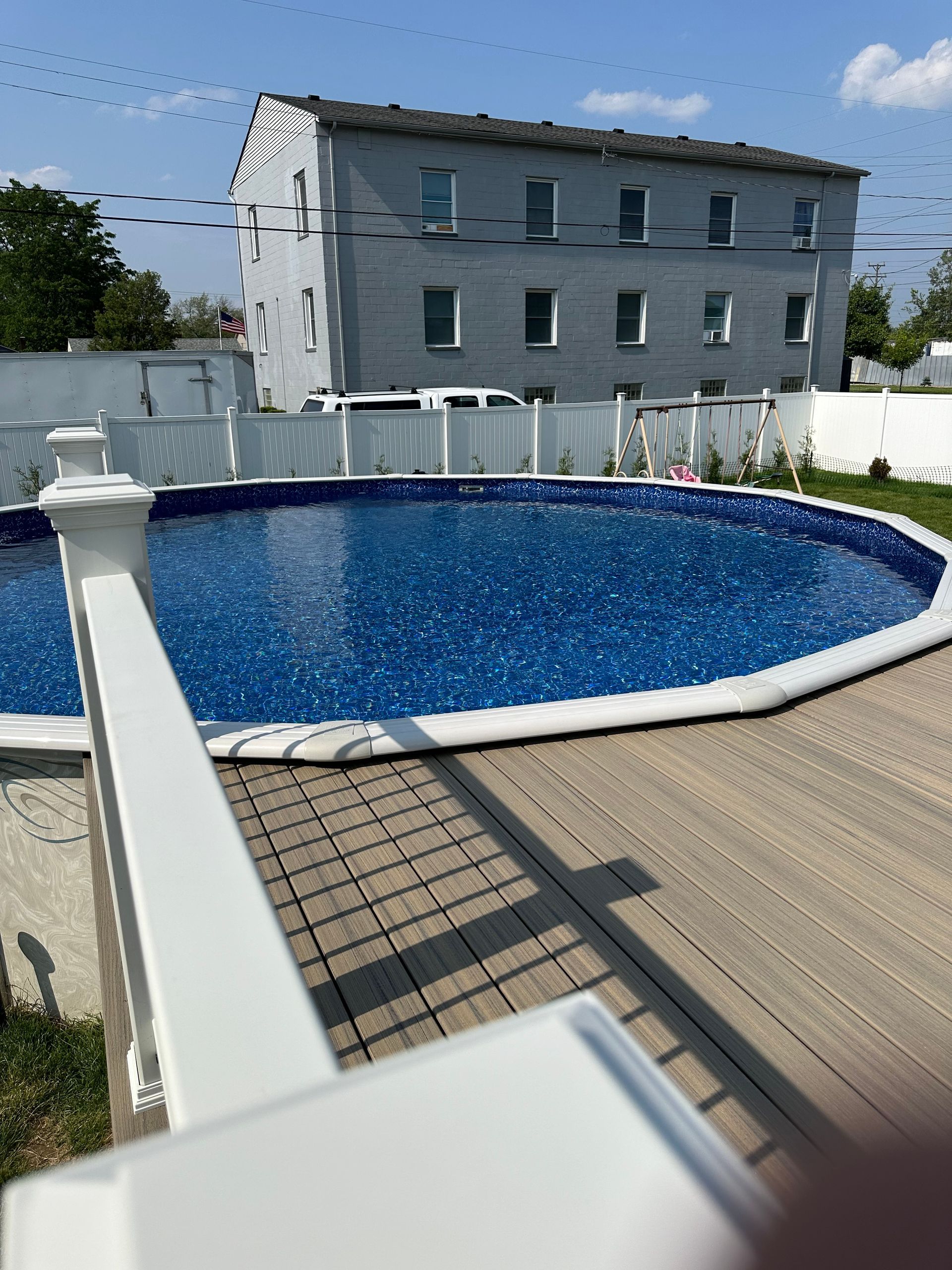 Above-ground pool with blue water and white trim, surrounded by a deck. A multi-story gray building is in the background.