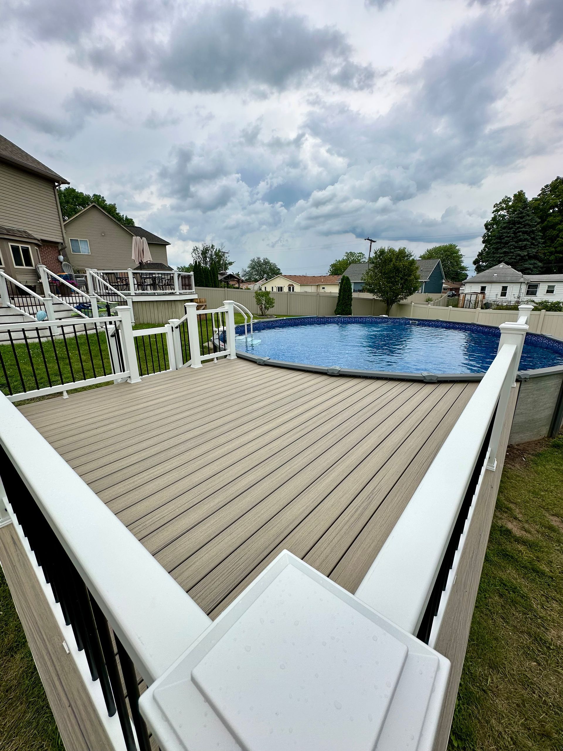 Deck with an above-ground pool, blue water, and overcast sky. Houses and green lawn surround it.