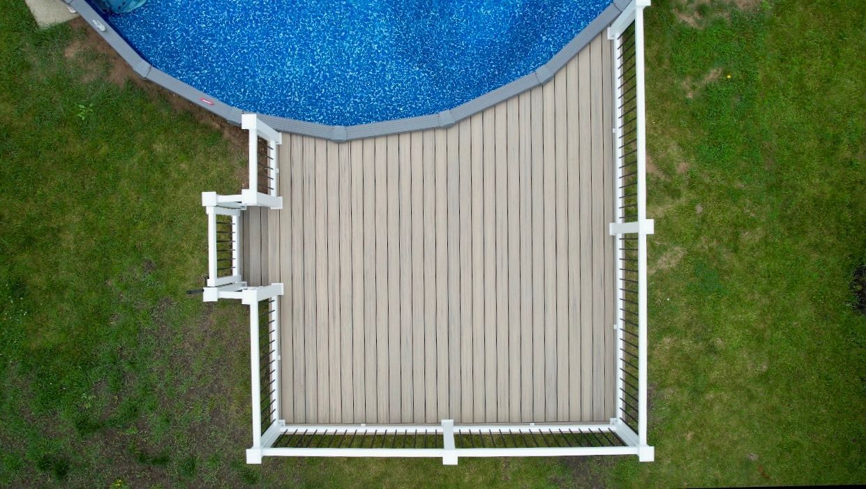 Overhead view of a wooden deck with a white railing next to a blue swimming pool in a grassy yard.