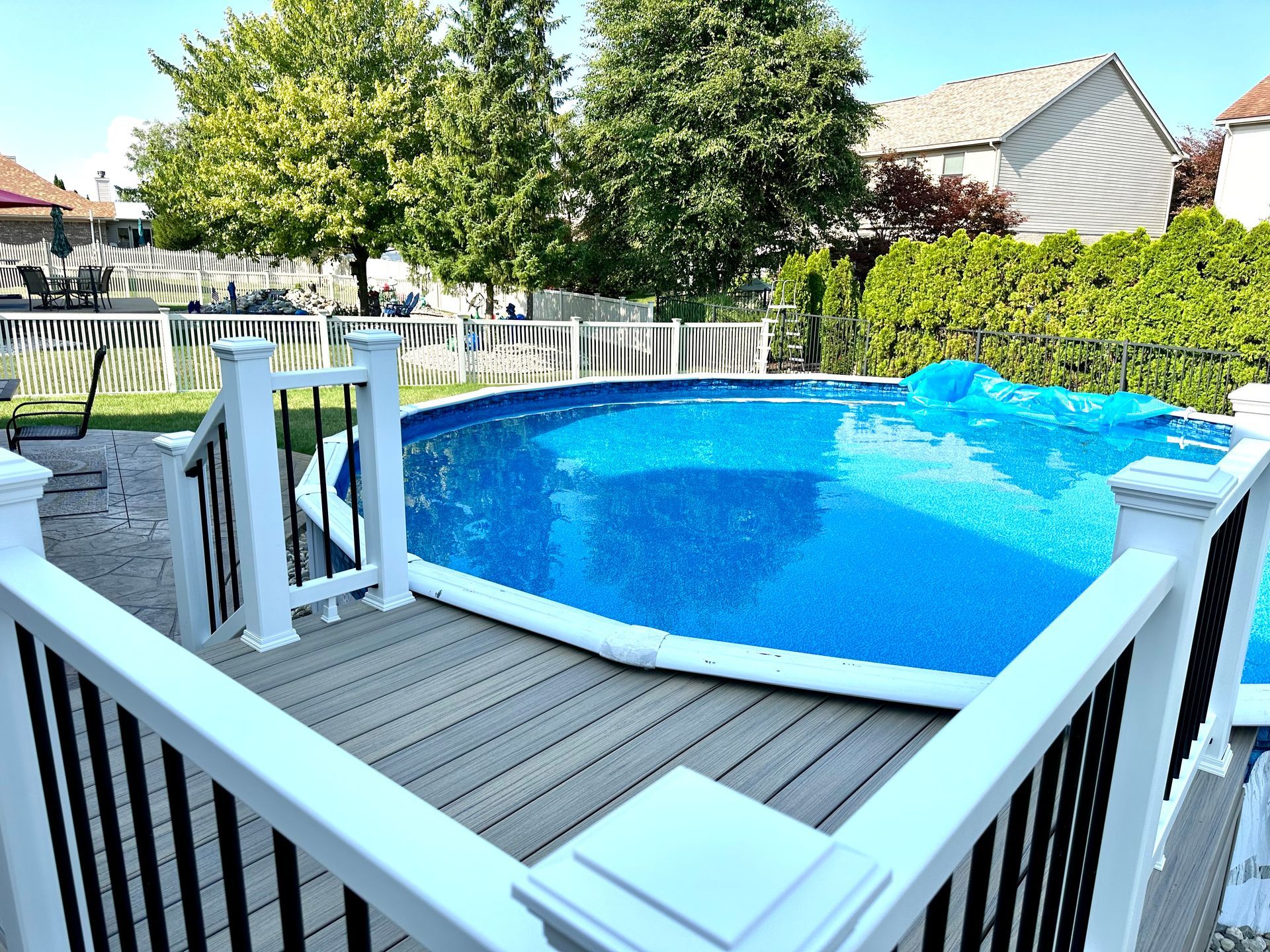 Above-ground pool with a wooden deck and white railing. Green trees and houses are visible in the background.