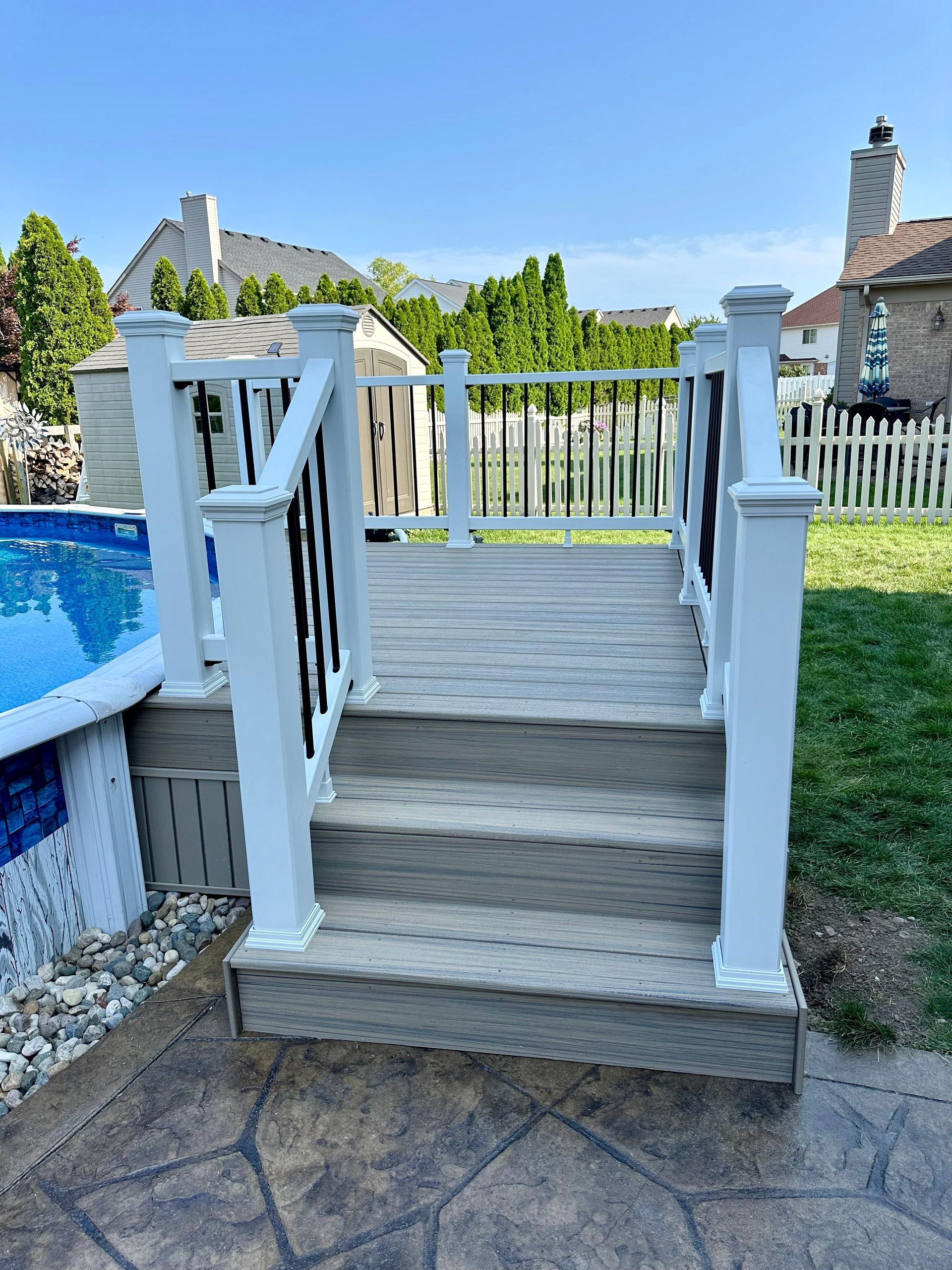 Deck with stairs leading to above-ground pool, white and black railings, sunny backyard.