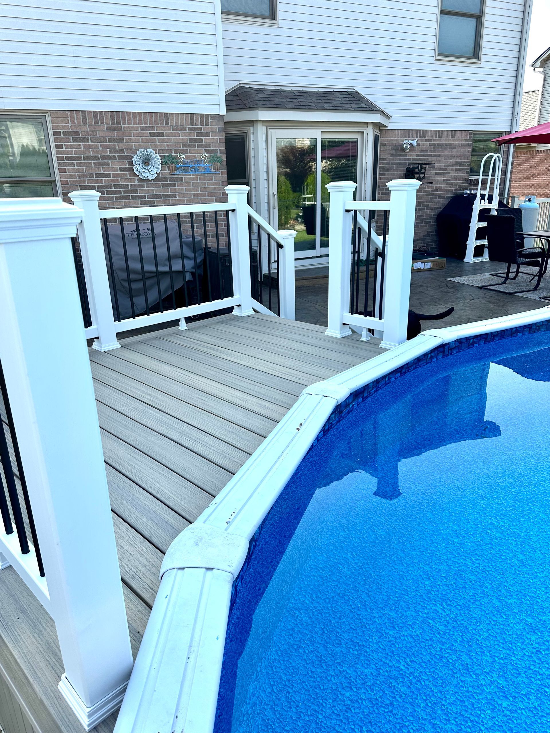 A deck with a white railing next to a blue swimming pool. Black railing supports. Brick house in the background.
