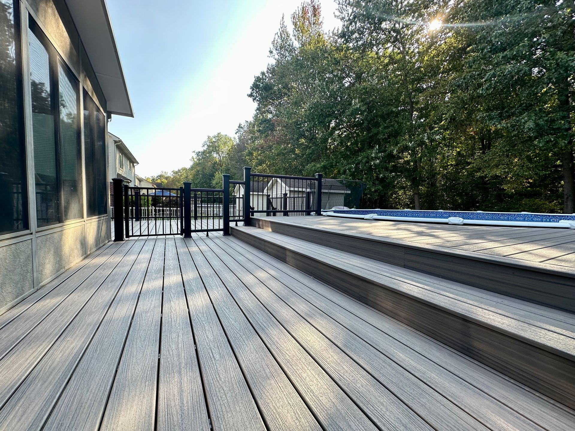 Wooden deck with steps leading to a pool, framed by a black fence, with sunlight shining through trees.
