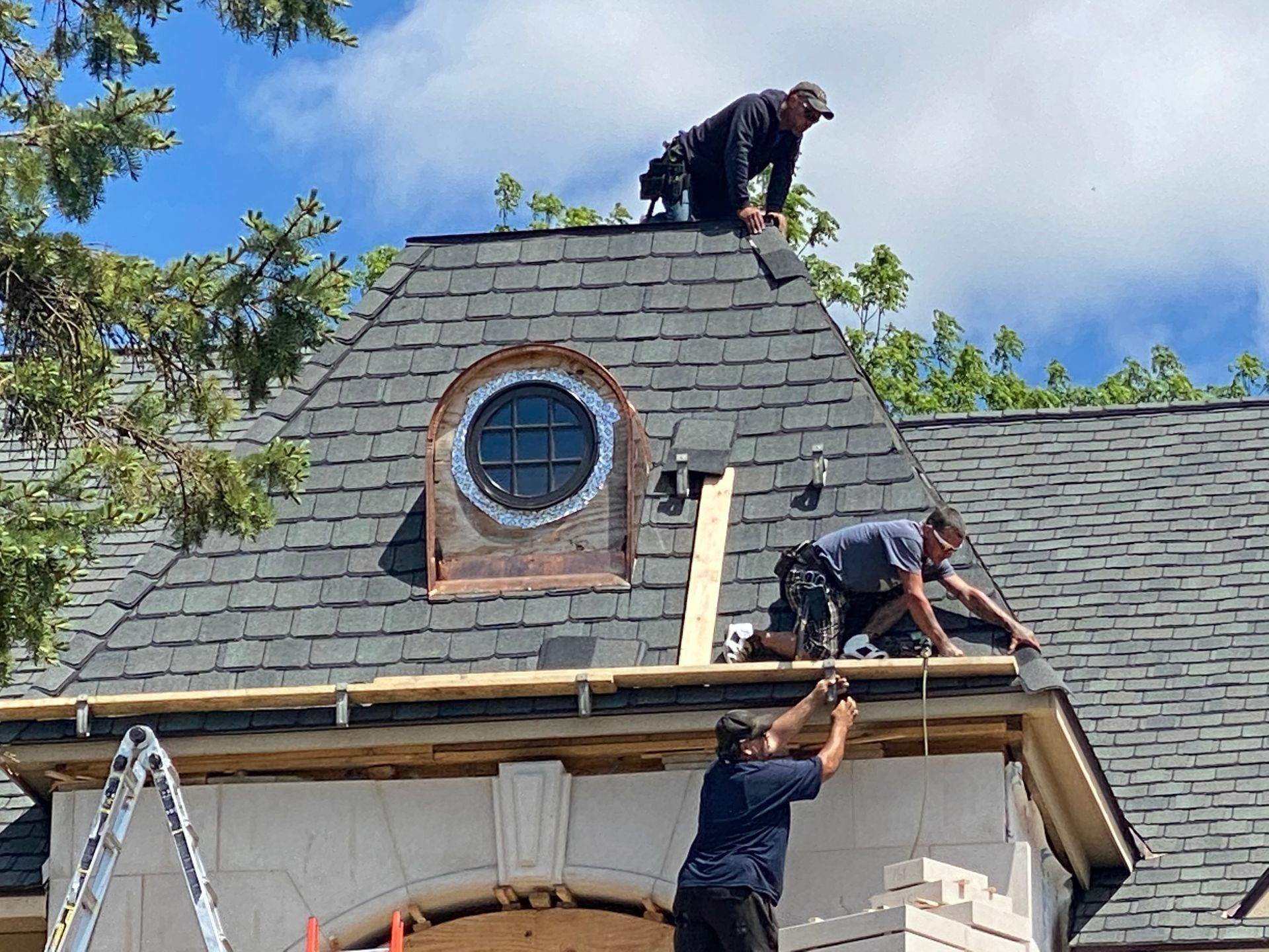 Three workers repairing a dark shingled roof on a building with arched window and a blue sky background.