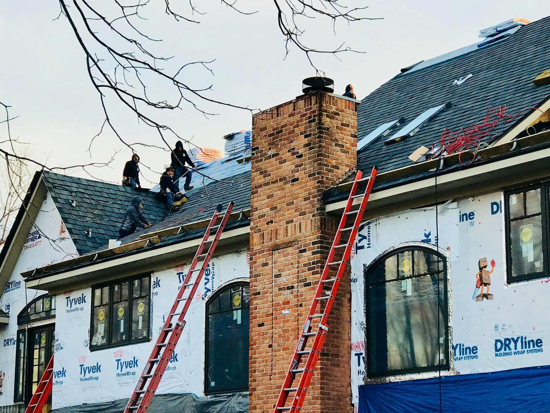 Construction workers on a roof, near a brick chimney. Ladders propped on the house. Blue tarp on the exterior.